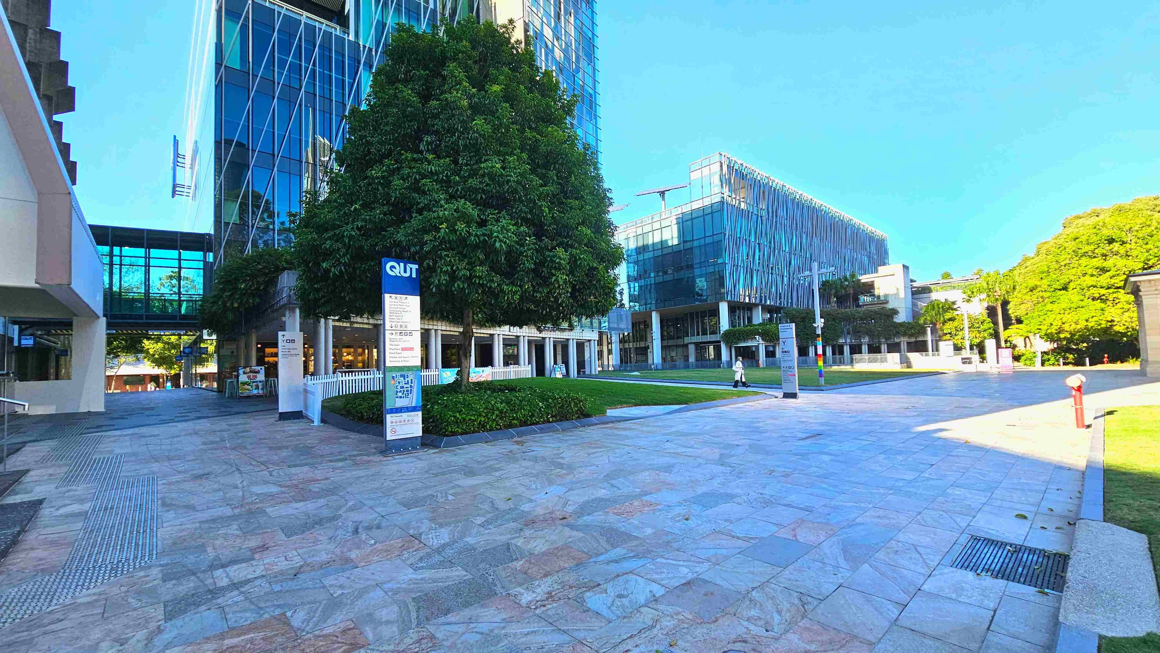A modern urban plaza with a wide stone-tiled surface, bordered by a large glass building with a crisscross pattern and a smaller building with a similar design. A prominent sign with 'QUT' stands near a lush green tree, surrounded by low hedges. A pedestrian walkway with tactile paving runs alongside. The area is open and spacious, with a few people walking and a red fire hydrant visible on the right.