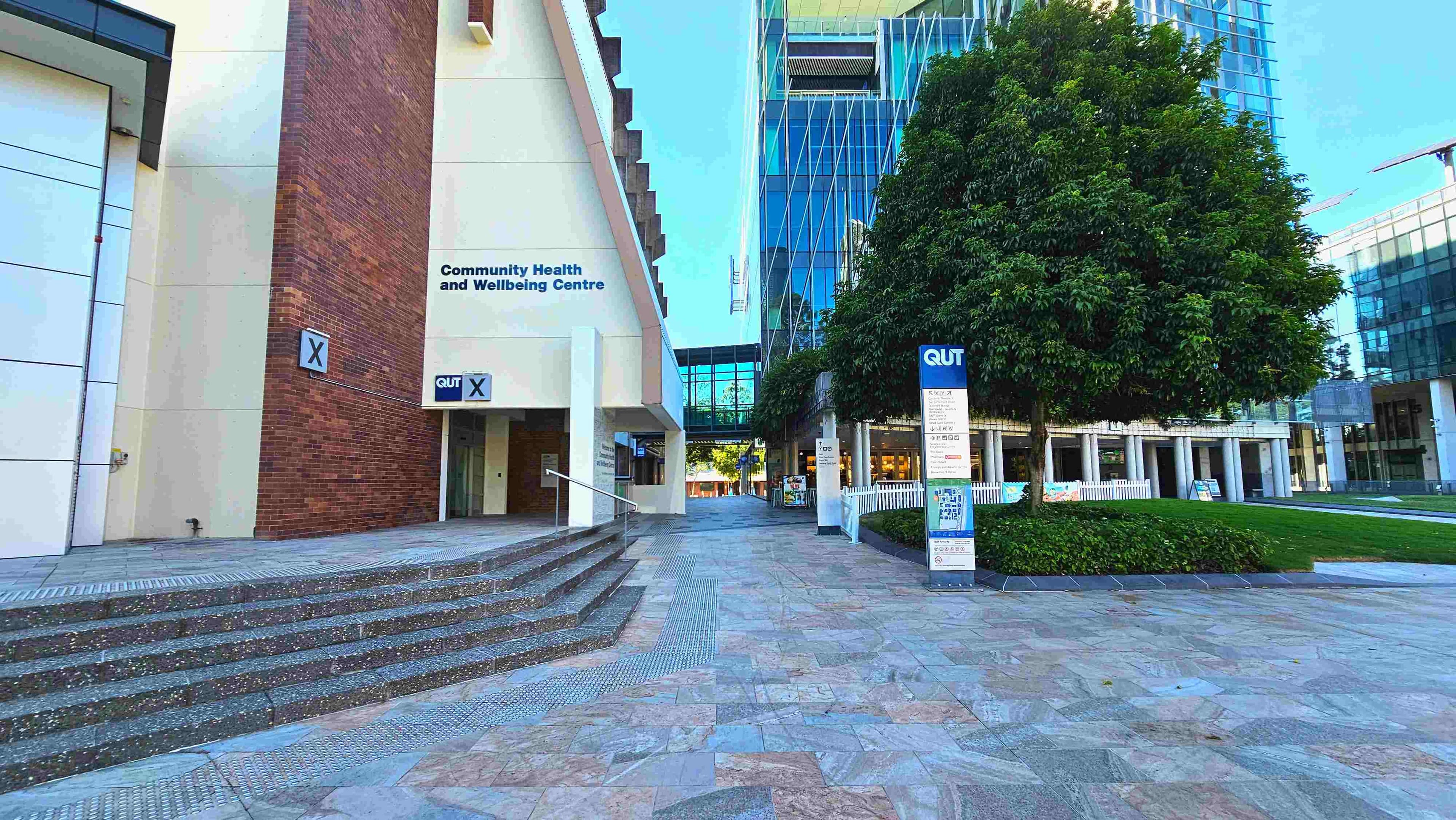A modern urban space featuring a Community Health and Wellbeing Centre with a brick and cream facade on the left. A set of wide, shallow steps leads up to the entrance. To the right, a large tree stands beside a signpost with 'QUT' branding. The ground is paved with patterned stone tiles. In the background, a glass-fronted building with a covered walkway connects the structures, creating a semi-enclosed area.