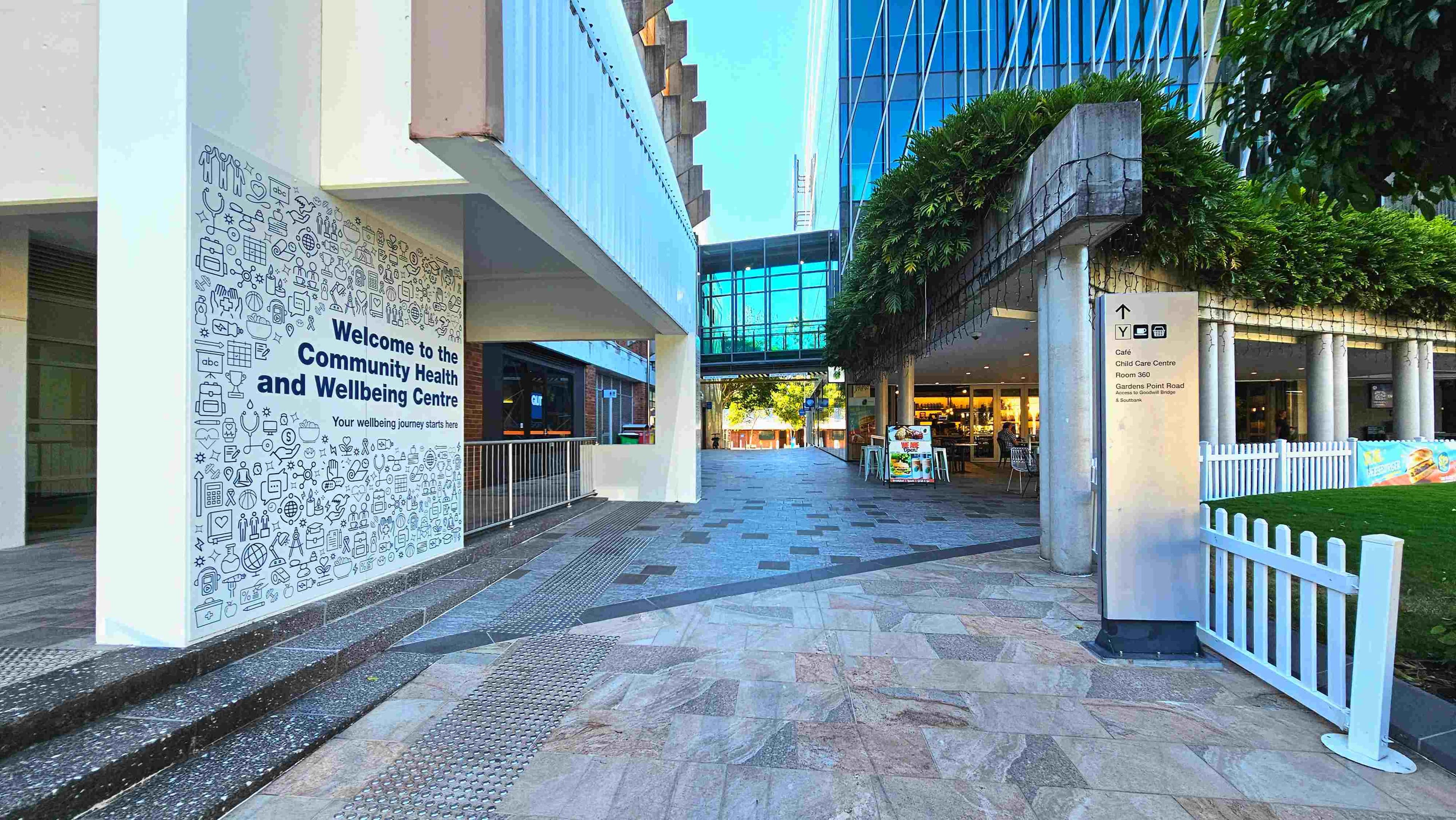 A modern outdoor walkway with a large sign reading 'Welcome to the Community Health and Wellbeing Centre' on a wall to the left. The path is paved with a mix of grey and pink tiles, leading towards a glass-walled building in the background. A white fence borders a grassy area on the right, and a tall signpost provides directions to various facilities. Greenery hangs from a concrete overhang, adding a touch of nature to the urban setting.