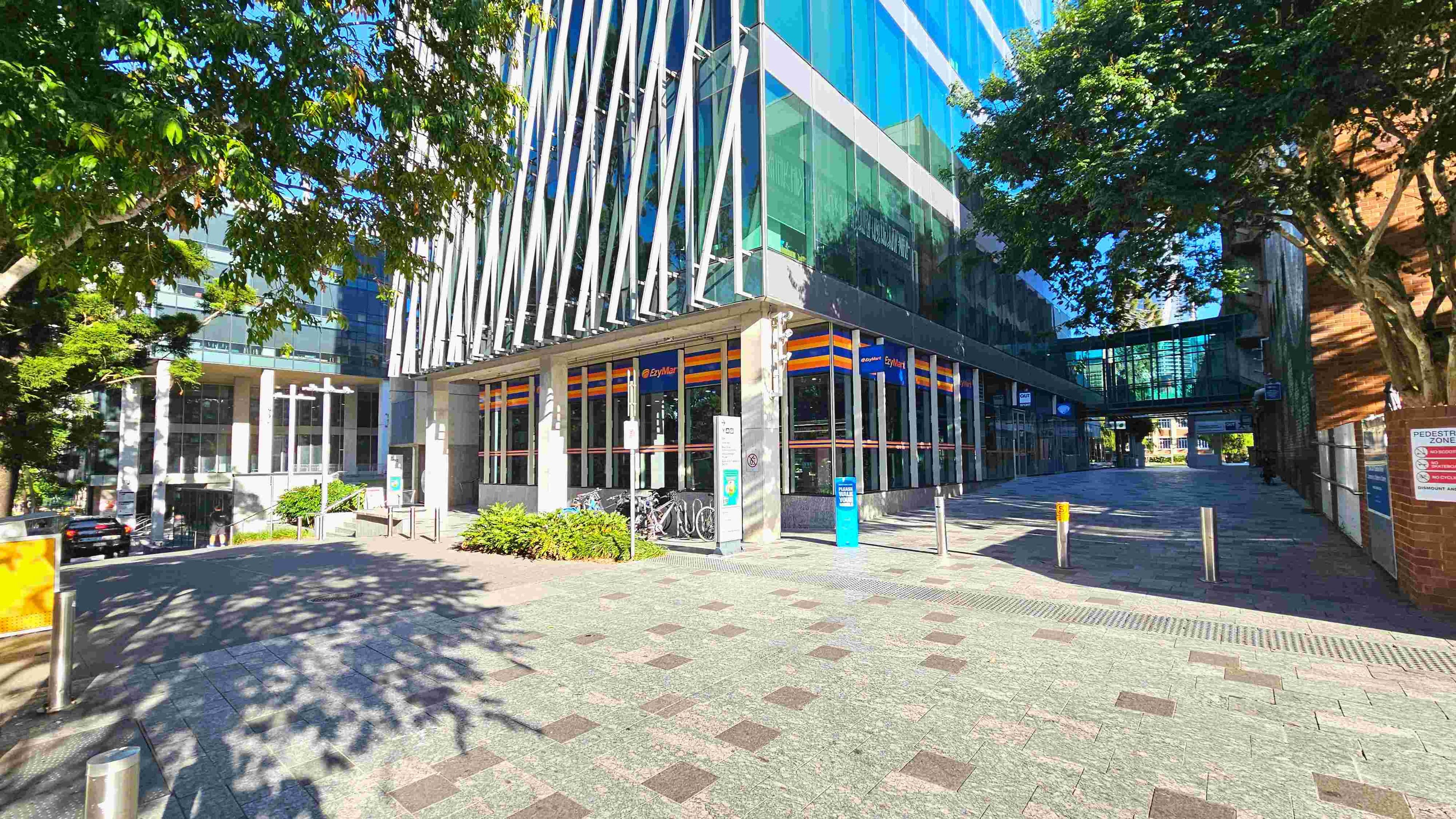 A modern urban scene featuring a glass-fronted building with diagonal white beams and a convenience store on the ground floor. The pavement is a mix of grey and brown tiles, leading to a pedestrian zone marked by bollards. A tree provides shade on the left, and a glass skybridge connects buildings in the background. Bicycles are parked near the entrance, and signage is visible on the walls.