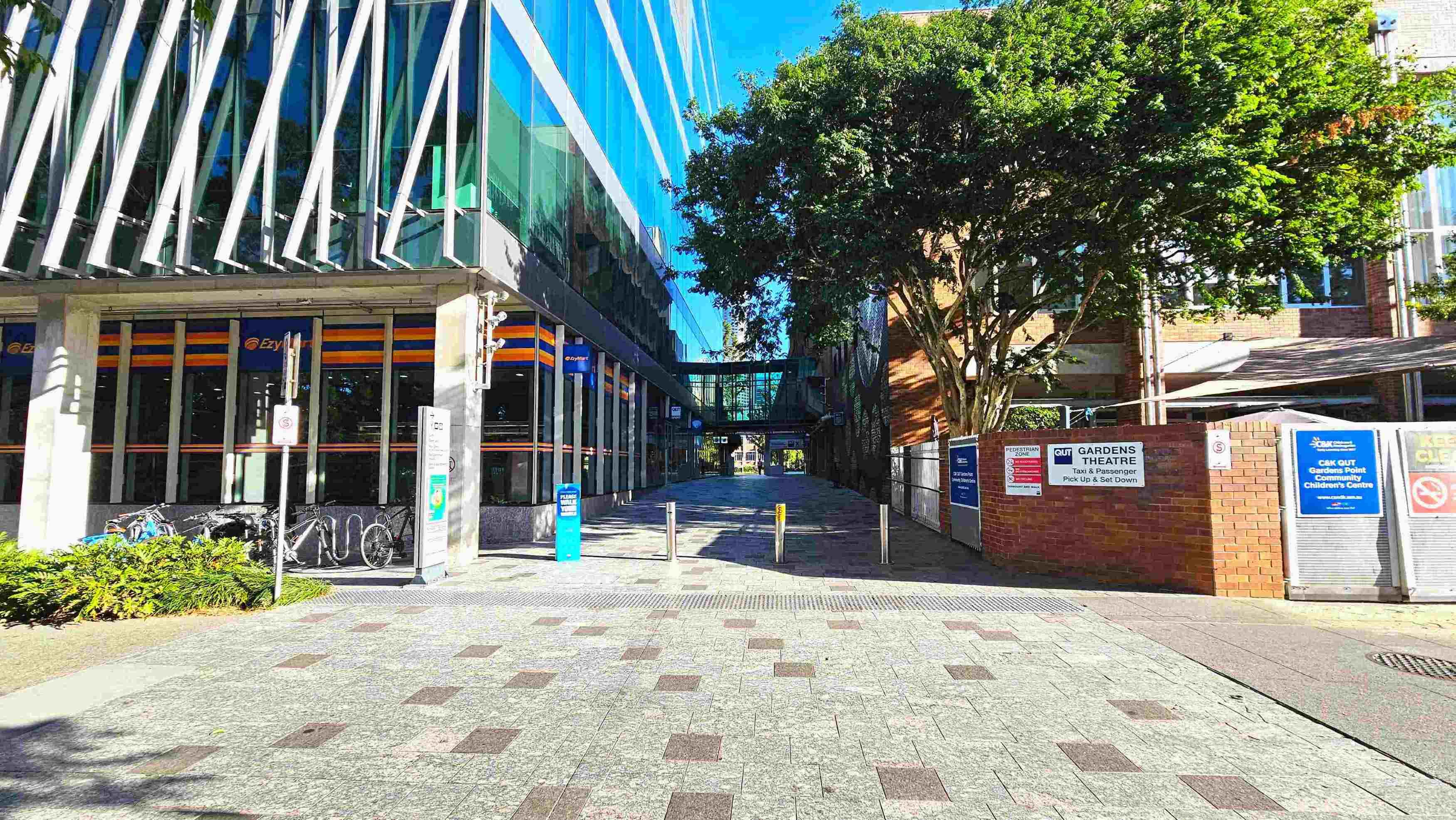 A modern urban walkway flanked by a glass-fronted building on the left and a brick building on the right. The glass building features diagonal white beams and a row of bicycles parked outside. A tree provides shade near the brick building, which has signage for Gardens Theatre. The path is paved with grey and brown tiles, leading to a glass bridge connecting the buildings. Bollards mark the entrance to the walkway.