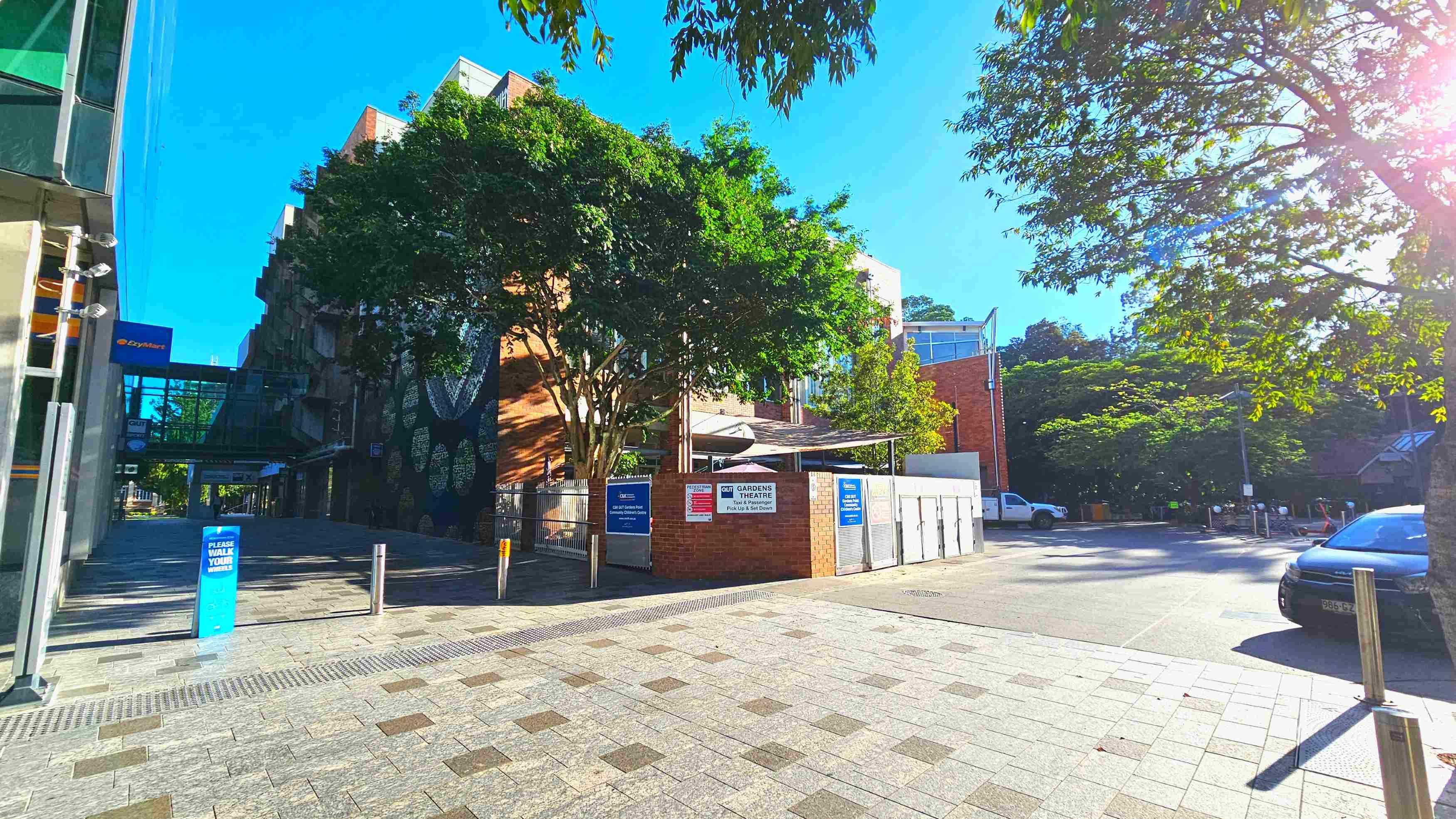 A paved area with a mix of light and dark grey tiles leads to a brick building partially obscured by lush green trees. Signs on the building indicate it is the QUT Gardens Theatre. A glass walkway connects to another structure on the left. Metal bollards line the path, and a parked car is visible on the right. The area feels open with trees providing shade and a clear view of the buildings.