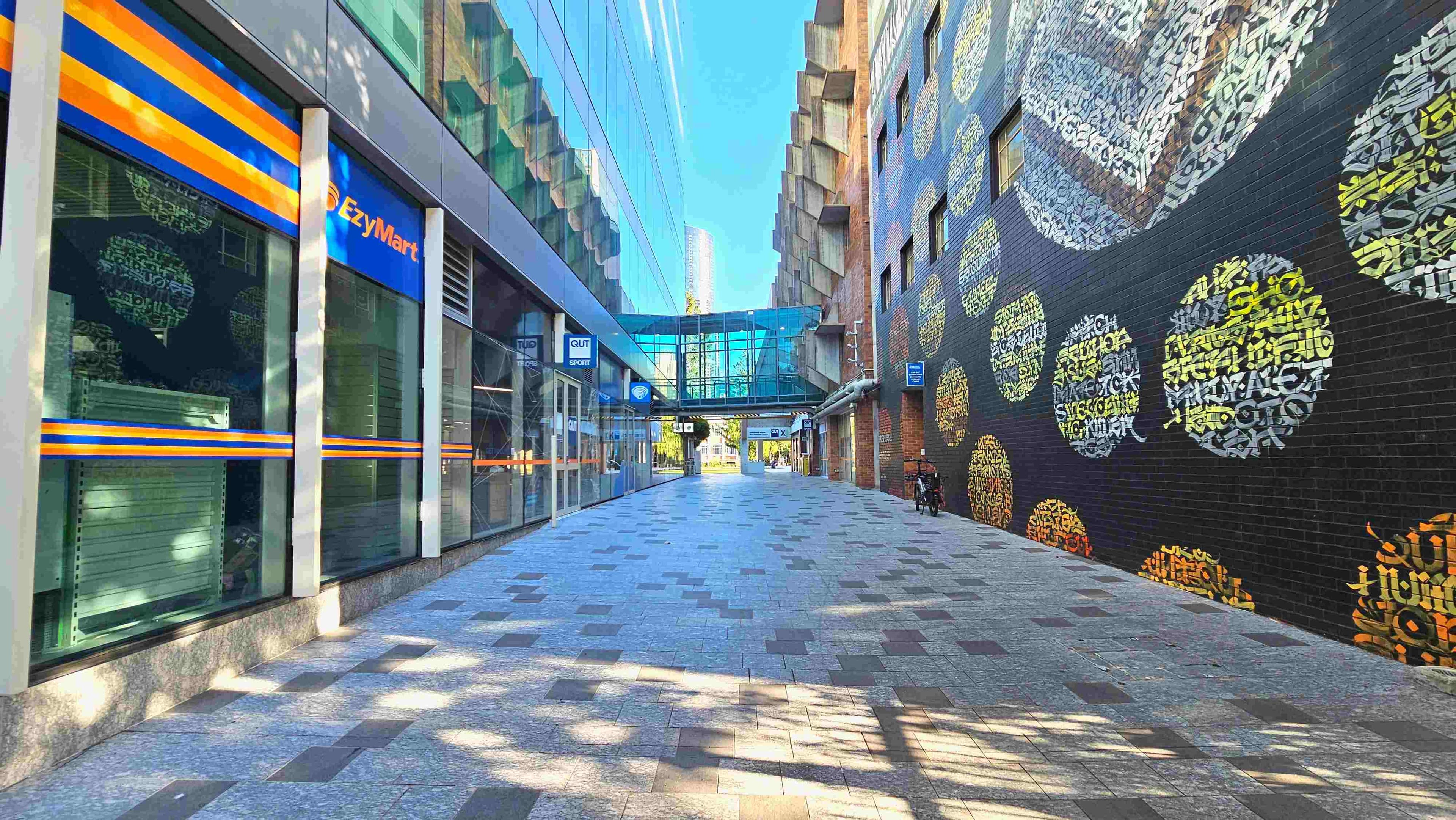 A pedestrian walkway flanked by a modern building with glass windows on the left and a brick wall adorned with colourful circular graffiti on the right. The path is paved with grey and brown tiles. A glass skybridge connects the buildings above. A bicycle is parked against the brick wall. Signs for 'EzyMart' and 'QUT Sport' are visible on the left. The area appears open and well-lit.