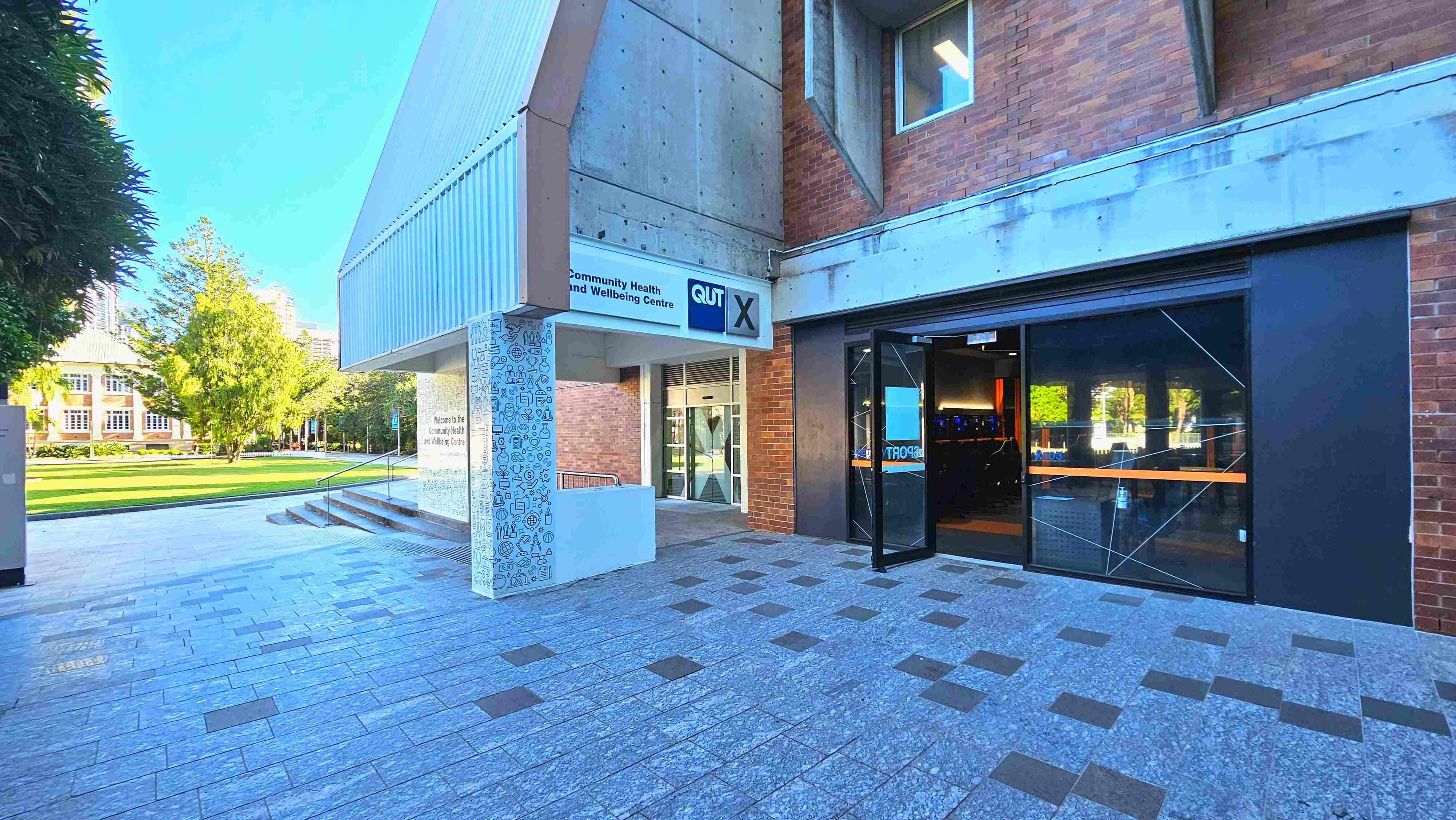 The image shows the entrance to a building with a sign reading 'Community Health and Wellbeing Centre' and 'QUT X'. The structure features a mix of brick and concrete with a modern design. A glass door is open, revealing a dimly lit interior. The ground is paved with grey tiles interspersed with darker squares. To the left, a grassy area with trees and a historic building is visible, suggesting an open campus environment.