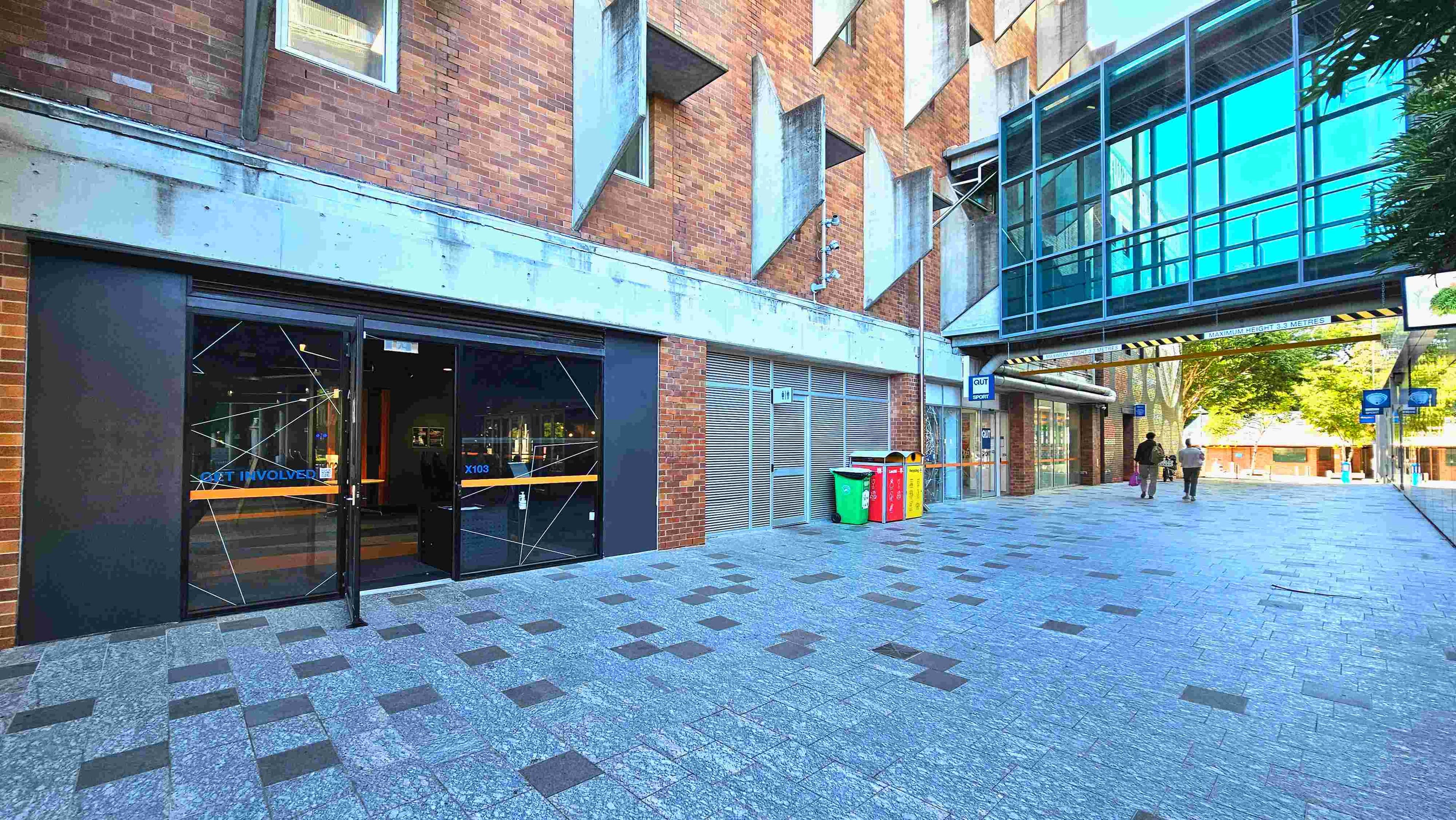 A modern urban walkway with a brick building on the left featuring geometric concrete protrusions. The building has a glass entrance with 'Get Involved' signage. Adjacent are colourful recycling bins. A glass skybridge connects to another structure, casting reflections on the ground. The walkway is paved with grey tiles interspersed with darker squares, leading to a tree-lined area in the distance.
