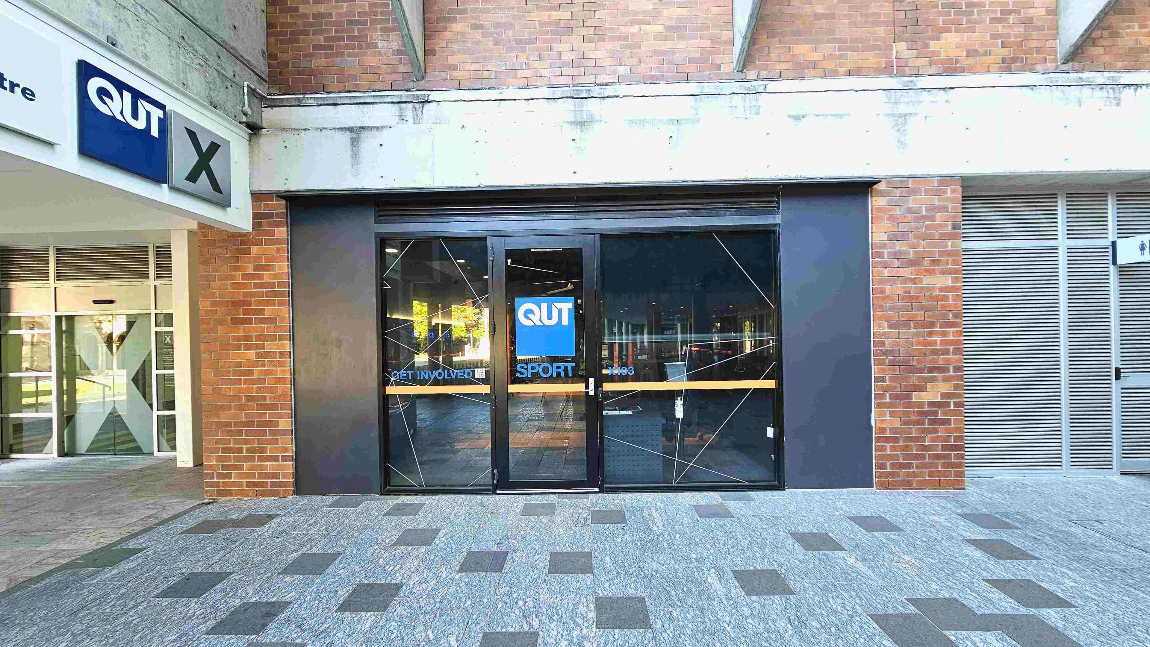 The image shows a brick building facade with a central glass entrance featuring the QUT Sport logo. The entrance has two large glass doors with geometric patterns and a blue QUT sign. To the left, a sign reads 'QUT X' above a glass door. The ground is paved with grey tiles in a checkered pattern. On the right, there is a louvred panel and a small sign indicating restrooms.