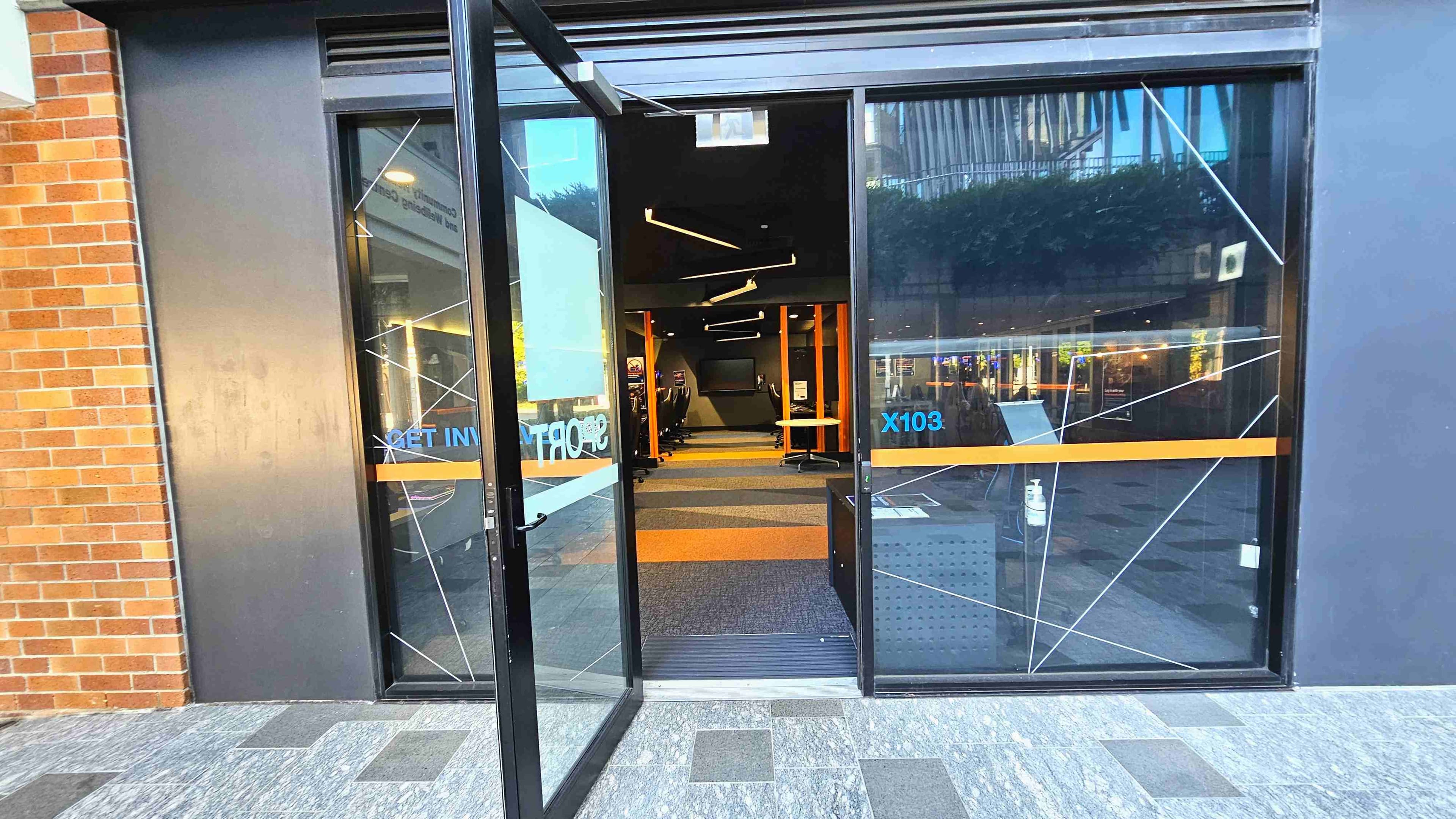 A modern entrance with large glass doors, one of which is open, revealing an interior with a patterned carpet and sleek black and orange furnishings. The exterior features a brick wall on the left and a dark grey panel on the right. The floor outside is tiled with grey and black squares. Inside, linear ceiling lights and a row of chairs are visible. A hand sanitiser dispenser is placed near the entrance.