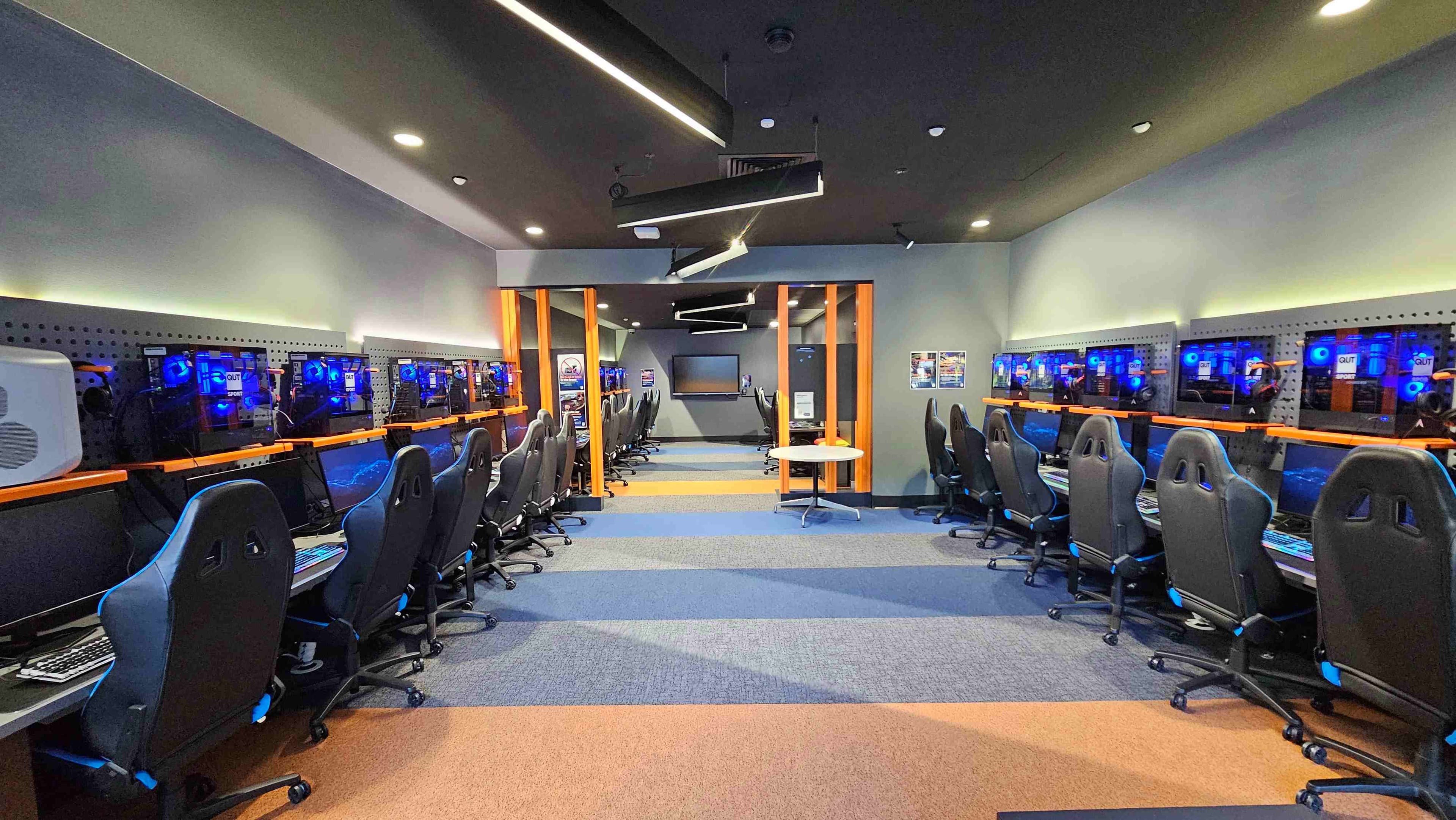 A modern gaming room with two rows of high-backed gaming chairs facing computer stations along both walls. Each station features a monitor and a glowing blue-lit computer tower mounted on a perforated panel. The room has a striped carpet in shades of blue and orange, and overhead, angular black light fixtures hang from the ceiling. At the far end, a large screen is mounted on the wall, flanked by orange vertical beams.