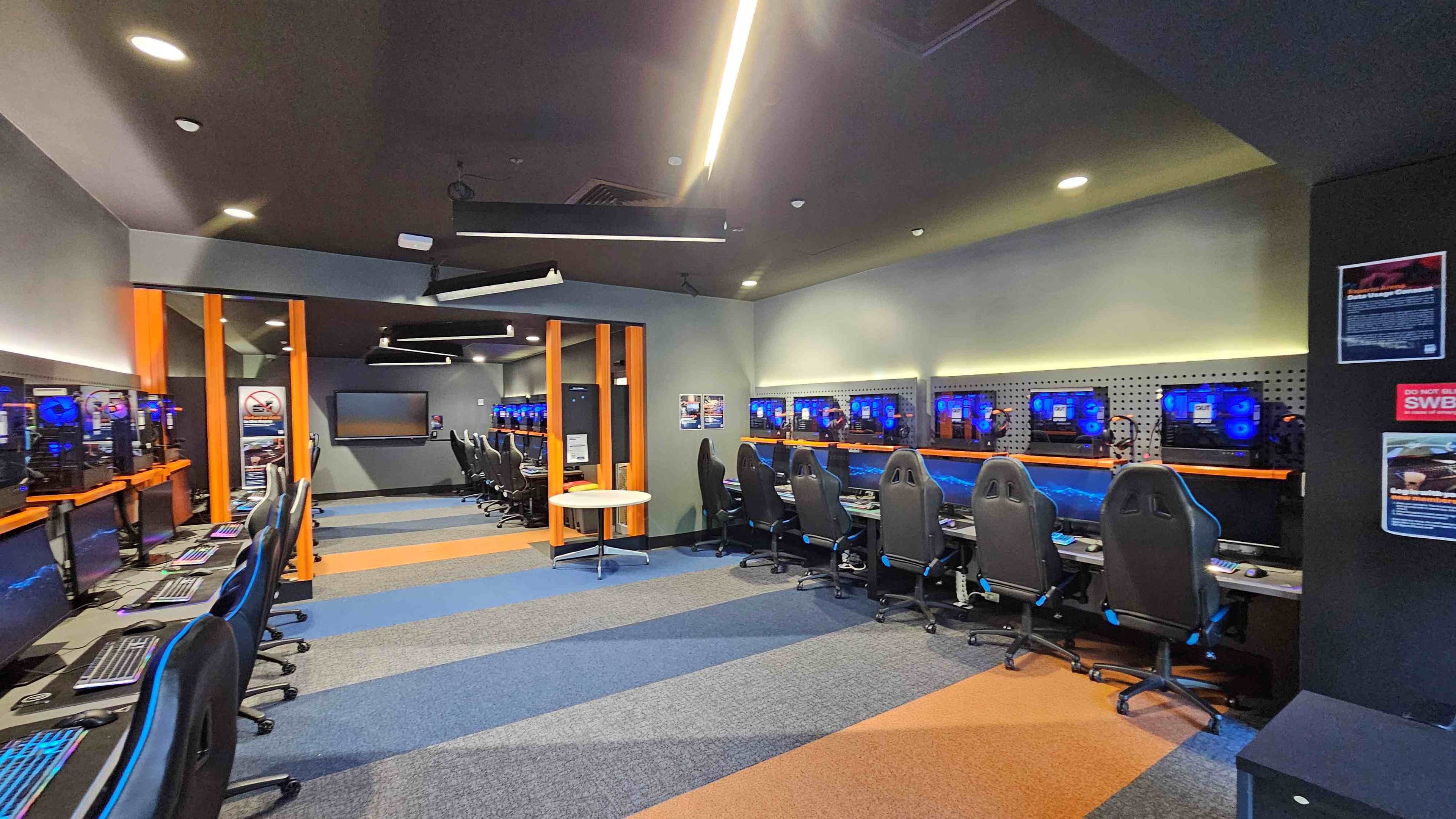 A modern gaming room with rows of high-backed gaming chairs facing computer stations. Each station has a monitor with blue LED lighting. The room features a mix of grey, blue, and orange carpet tiles, creating a vibrant pattern. Orange vertical beams divide the space, and posters are visible on the walls. The ceiling has recessed lighting and angular fixtures, adding a futuristic touch.