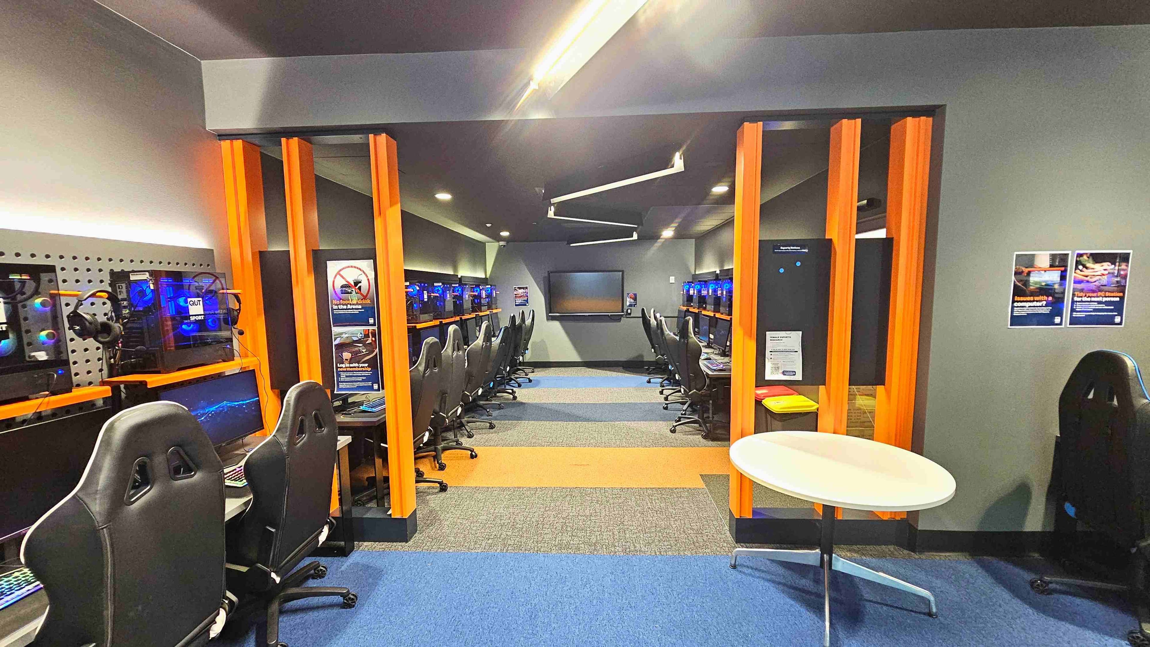 A modern gaming room with rows of high-backed black chairs facing computer stations on both sides. Each station has a monitor, keyboard, and headset, with blue-lit computer towers visible. The room is divided by orange vertical beams and features a grey wall with a large screen at the far end. The floor is carpeted in blue, orange, and grey sections. Posters are on the walls, and a small round table is in the foreground.