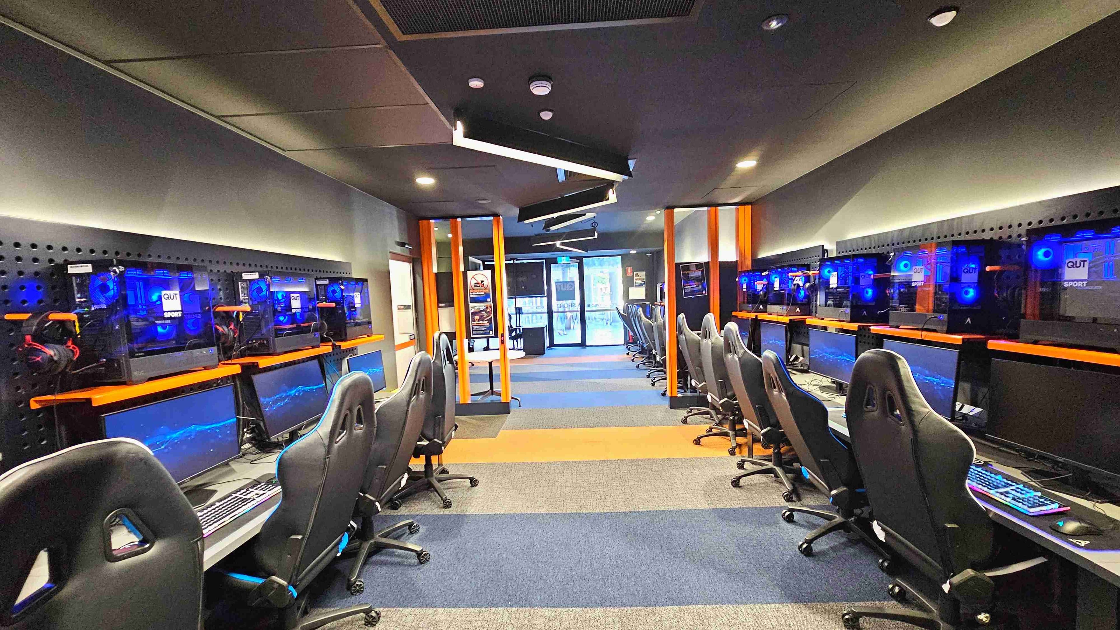 A modern gaming room with two rows of high-backed gaming chairs facing computer stations. Each station has a monitor, keyboard, and a glowing blue-lit CPU. The room features a dark ceiling with angular light fixtures and orange vertical beams. The floor is carpeted with alternating blue and orange stripes. At the far end, glass doors lead to another area, flanked by posters and signage.