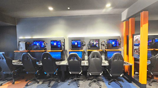 A row of five gaming stations is set against a wall with pegboard panels. Each station features a desktop computer with illuminated components, a monitor, keyboard, and headphones. Black gaming chairs are positioned in front of each setup. The floor is carpeted with a mix of grey and blue tiles. Bright orange vertical beams frame the right side, where a sign prohibits food and drinks. Ceiling lights provide illumination.