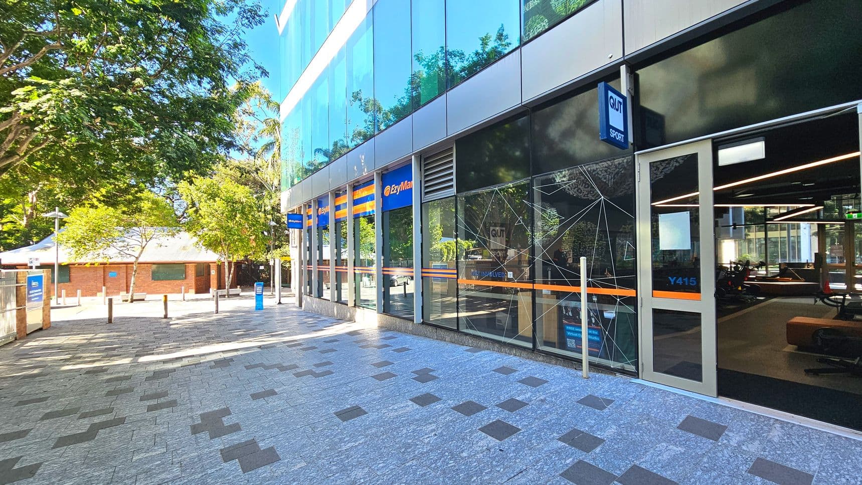 A modern building with large glass windows and a sign reading 'QUT Sport' above a door marked 'Y415'. The building's facade features blue and orange signage for 'EzyMart'. The pavement is a mix of grey and dark tiles, leading to a tree-lined area with bollards. A brick building with a white roof is visible in the background. Inside, gym equipment is visible through the glass.