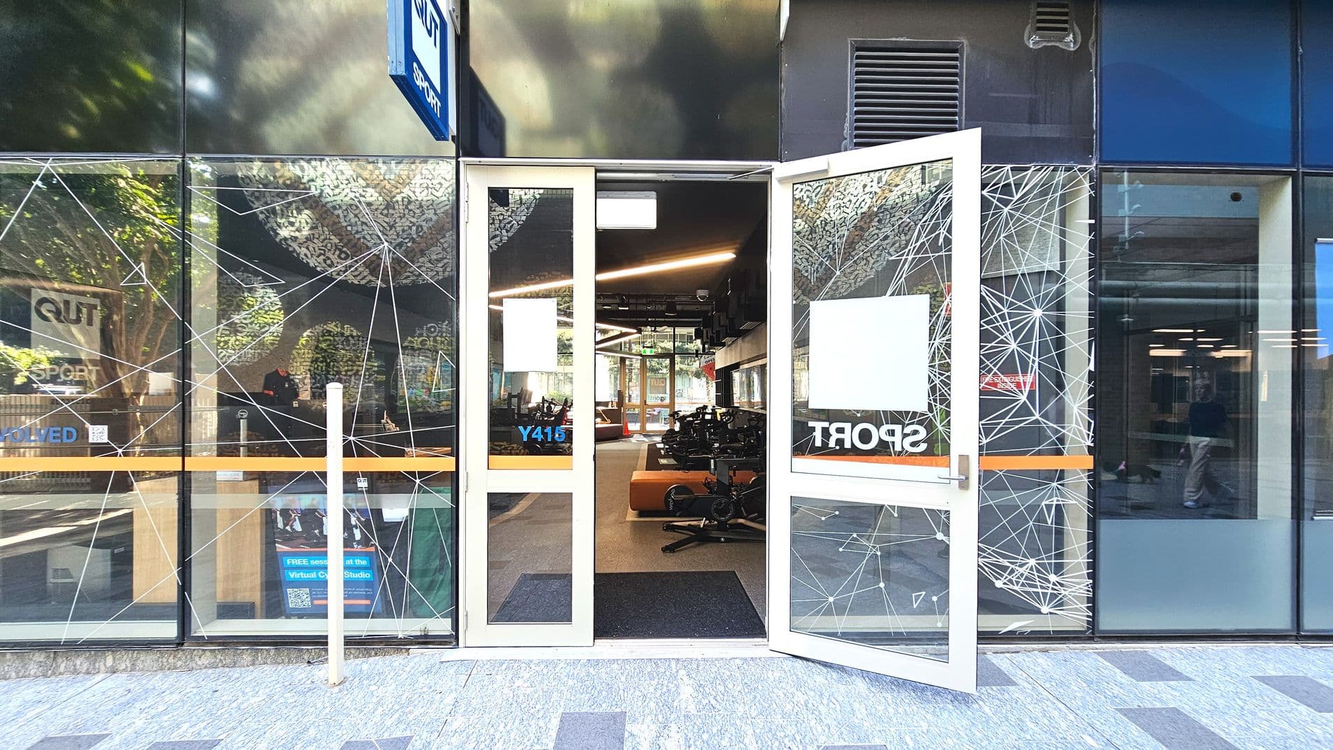 A modern entrance with glass doors open wide, revealing an interior with exercise equipment. The glass facade features geometric patterns and signage for 'QUT Sport'. The floor outside is tiled in grey, transitioning to a dark mat inside. A sign with 'Y415' is visible on the door. The space inside appears open with visible lighting fixtures and a reflective ceiling.