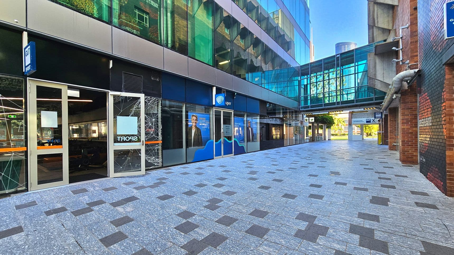 A modern urban walkway with a tiled floor featuring a pattern of grey and dark squares. On the left, a building with large glass windows displays signage for a sports facility. Above, a glass-enclosed pedestrian bridge connects two buildings. The right side has a brick wall with visible piping. In the distance, a green area is visible through the open end of the walkway.
