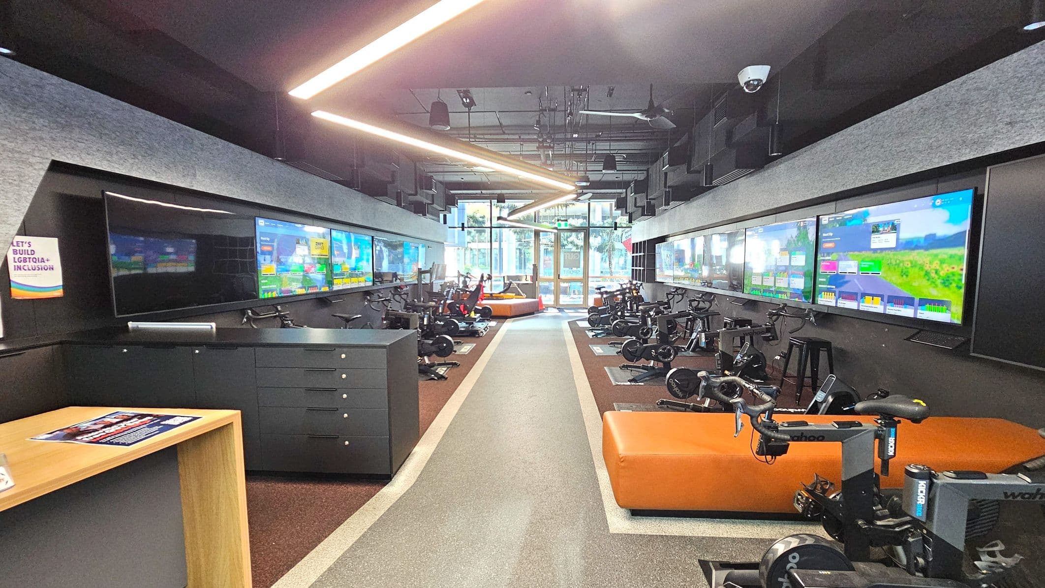 A modern indoor cycling studio with a central aisle flanked by stationary bikes on either side. Large screens displaying virtual cycling routes are mounted on the walls above the bikes. The floor is a mix of grey and brown materials, with a distinct central pathway. The ceiling features linear lighting and acoustic panels. At the far end, glass doors lead outside, and orange seating is visible near the entrance.