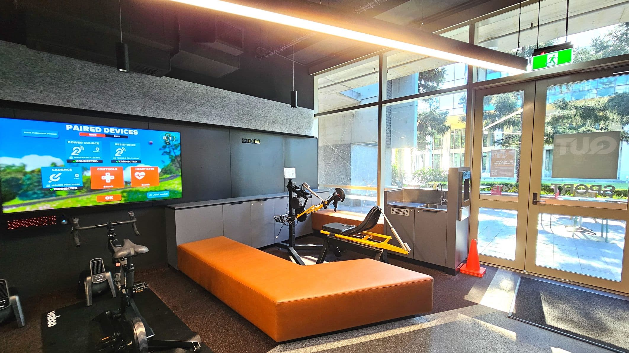 A modern indoor cycling area features a large screen displaying paired device information, mounted on a grey wall. Below, a stationary bike is positioned on a black mat. An L-shaped orange bench surrounds a yellow exercise machine. To the right, glass doors with an exit sign lead outside, flanked by a water dispenser and an orange cone. The floor transitions from dark carpet to a lighter mat near the entrance.