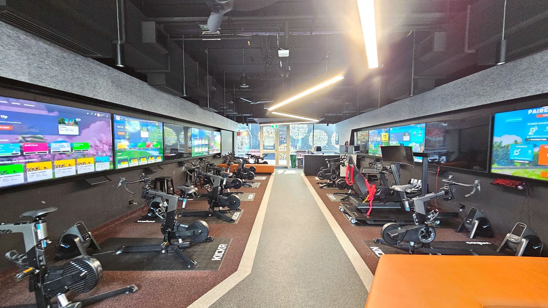 A modern indoor cycling studio with rows of stationary bikes on either side of a central aisle. Large digital screens line the walls above the bikes, displaying colourful graphics and information. The floor is carpeted with a central grey strip flanked by brown sections. The ceiling is high with exposed ductwork and linear lighting. At the far end, glass doors lead to another area, with decorative patterns visible on the glass.