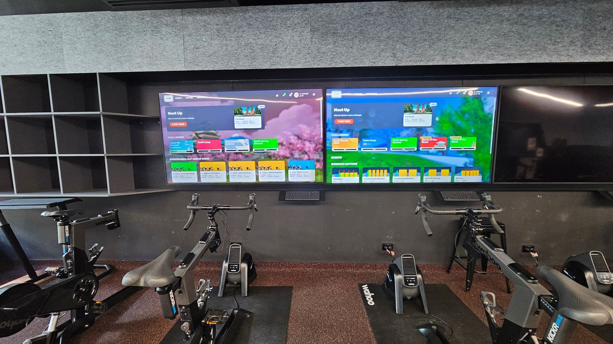 A modern indoor cycling studio featuring two large digital screens displaying colourful workout interfaces. Below the screens are three stationary bikes with sleek black frames and padded seats, positioned on dark mats. To the left, there is a wall-mounted shelving unit with multiple open compartments. The floor is covered in a speckled dark carpet, and the walls are lined with grey acoustic panels.