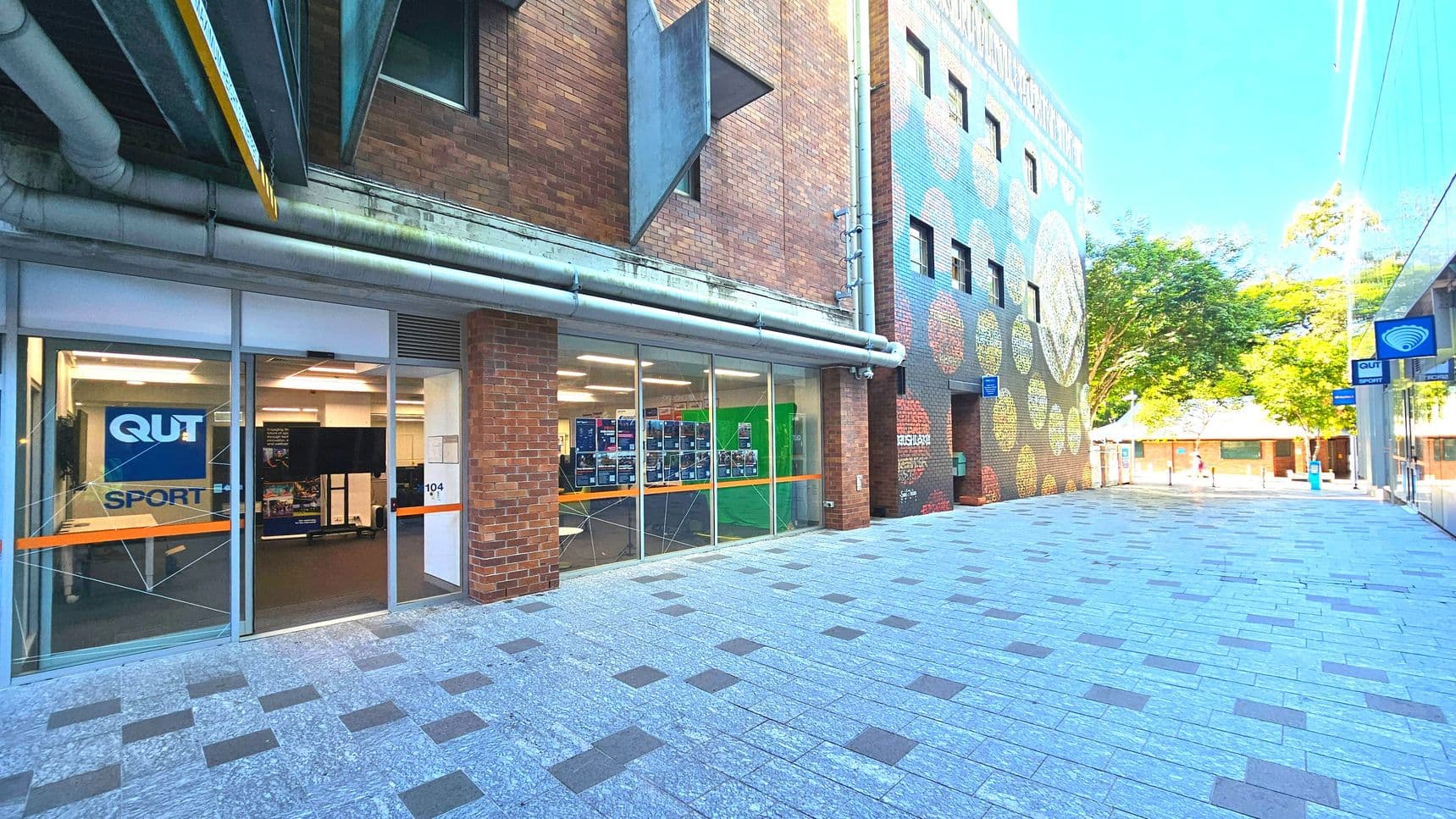 A brick building with large glass windows displays a sign reading 'QUT Sport'. The entrance is framed by a brick column. Adjacent is a colourful mural on another brick wall with circular patterns. The ground is paved with light grey tiles interspersed with darker squares. A blue sign with a Wi-Fi symbol hangs above. Trees and a small building are visible in the background, creating an open courtyard feel.
