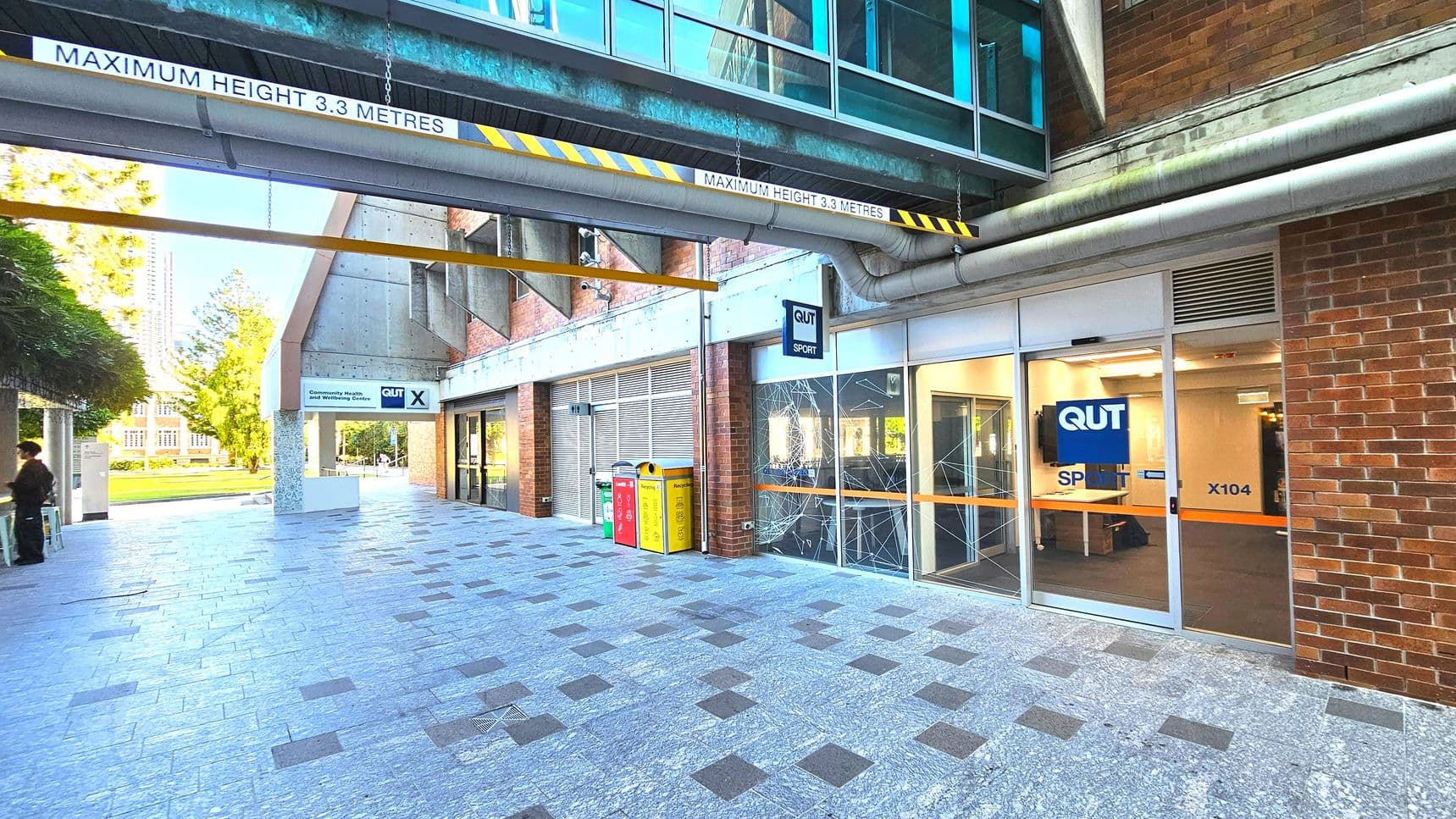 A covered walkway with a maximum height sign of 3.3 metres is visible, featuring a glass and metal overpass above. The ground is paved with grey and dark square tiles. On the right, a brick building with glass doors marked 'QUT Sport X104' is present. To the left, a sign for 'QUT' and bins in red, yellow, and green are seen. The area feels open with a view of a grassy area and trees in the background.