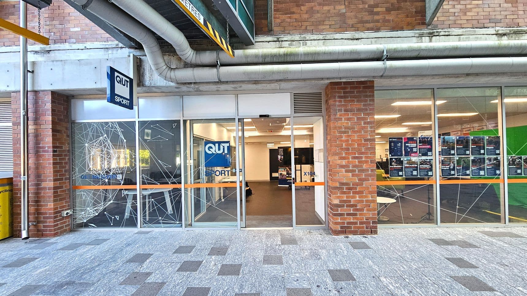 A brick building facade with large glass windows and a central entrance. The windows are decorated with white geometric patterns and an orange stripe. A sign reads 'QUT Sport' above the entrance. Inside, a well-lit room with posters on the walls is visible. The floor outside is a mix of grey and brown tiles. Overhead, large pipes run along the building, and a yellow clearance sign hangs above.