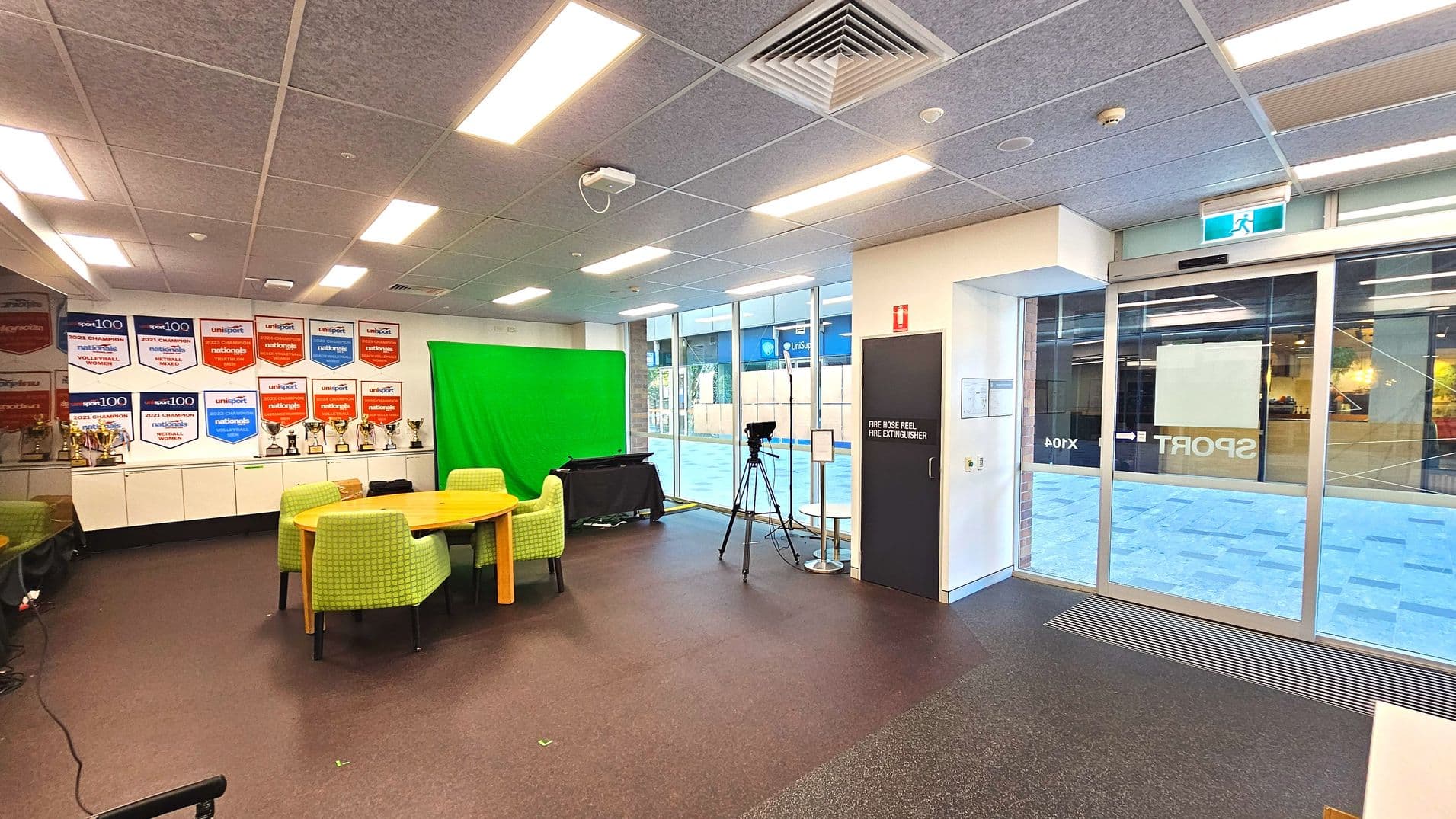 The room features a wall with numerous sports banners and trophies on white cabinets. A green screen is set up beside a round wooden table surrounded by green upholstered chairs. A camera on a tripod is positioned near a glass sliding door, which has a visible exit sign above. The floor is carpeted, and the ceiling has recessed lighting and ventilation panels. A door marked 'Fire Hose Reel Fire Extinguisher' is on the right.