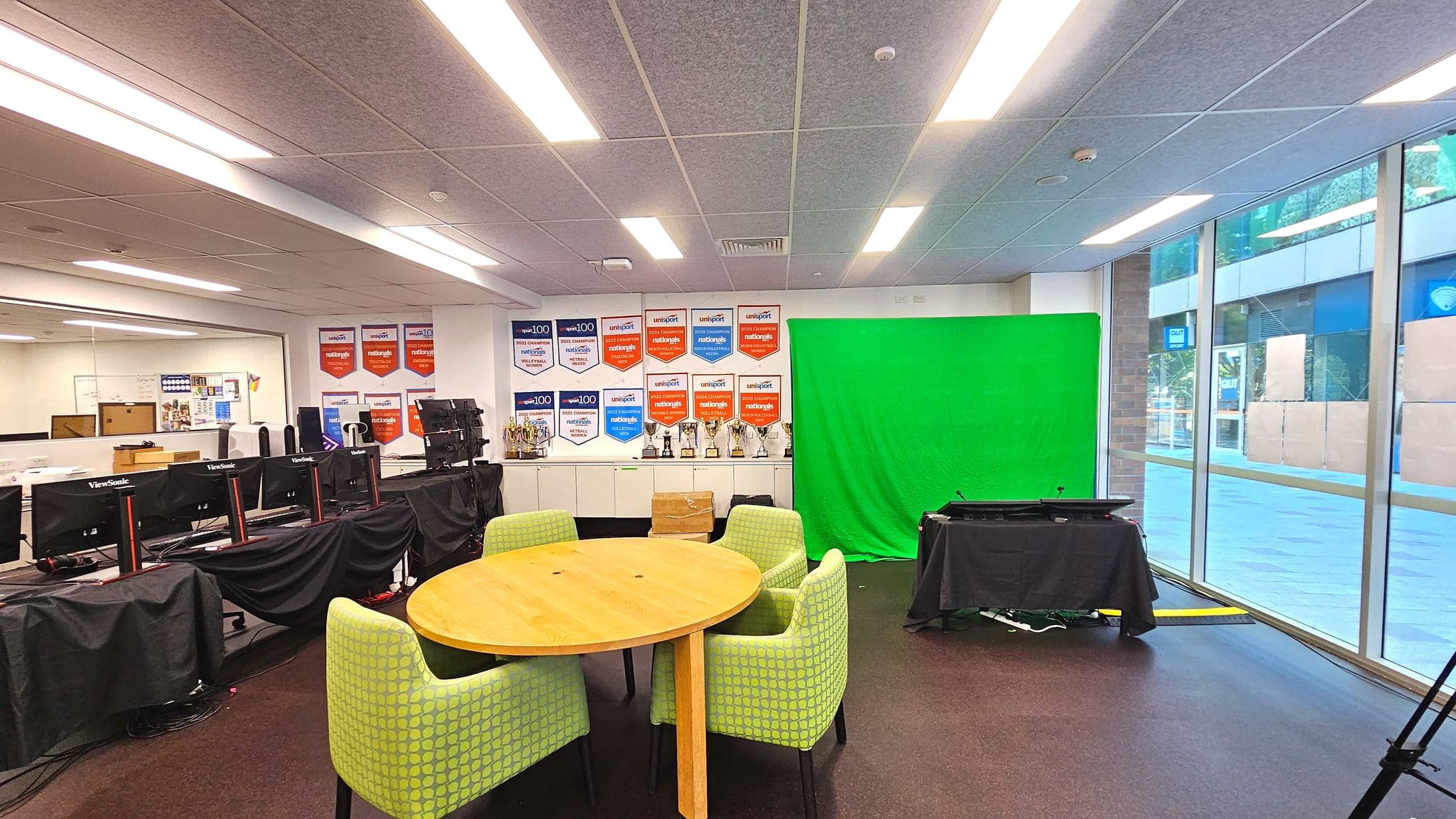An indoor space featuring a round wooden table surrounded by four green upholstered chairs with a geometric pattern. To the right, a large green screen is set up against a wall adorned with numerous award banners and trophies on a shelf. Several computer monitors are covered with black cloths on the left. The room has a low ceiling with fluorescent lighting and large glass windows on the right, offering a view of an outdoor area.