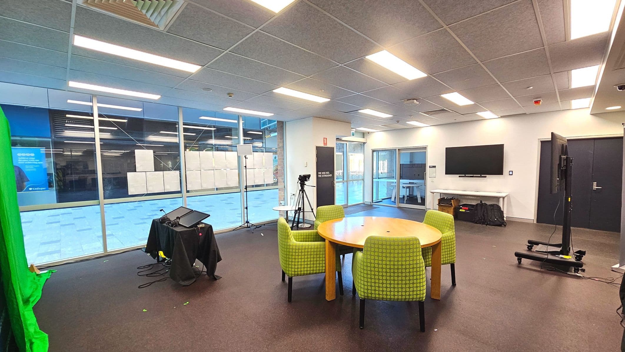 A modern room with a large glass wall on the left, featuring a green screen setup and a camera on a tripod. In the centre, a round wooden table is surrounded by four green upholstered chairs. A large flat-screen TV is mounted on the right wall above a white console table. The floor is carpeted in dark grey, and the ceiling has recessed lighting. A set of double glass doors is visible in the background.
