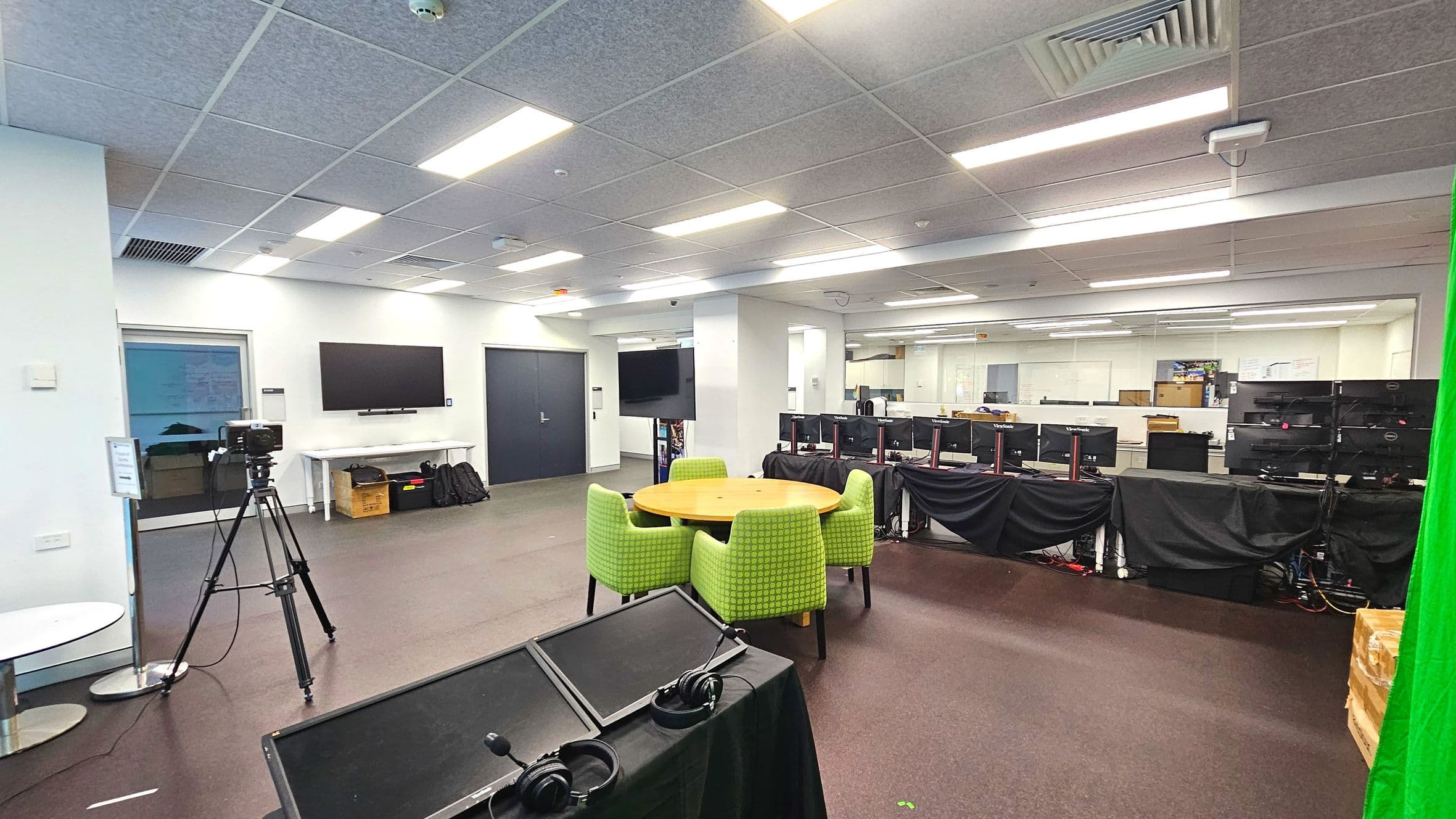 A spacious room with a modern office layout. In the foreground, a round wooden table is surrounded by four green upholstered chairs. To the left, a camera on a tripod faces a wall-mounted screen above a white table with bags underneath. Several computer monitors are lined up on desks covered with black cloths. The ceiling features recessed lighting and air vents. The floor is carpeted, and a green screen is partially visible on the right.