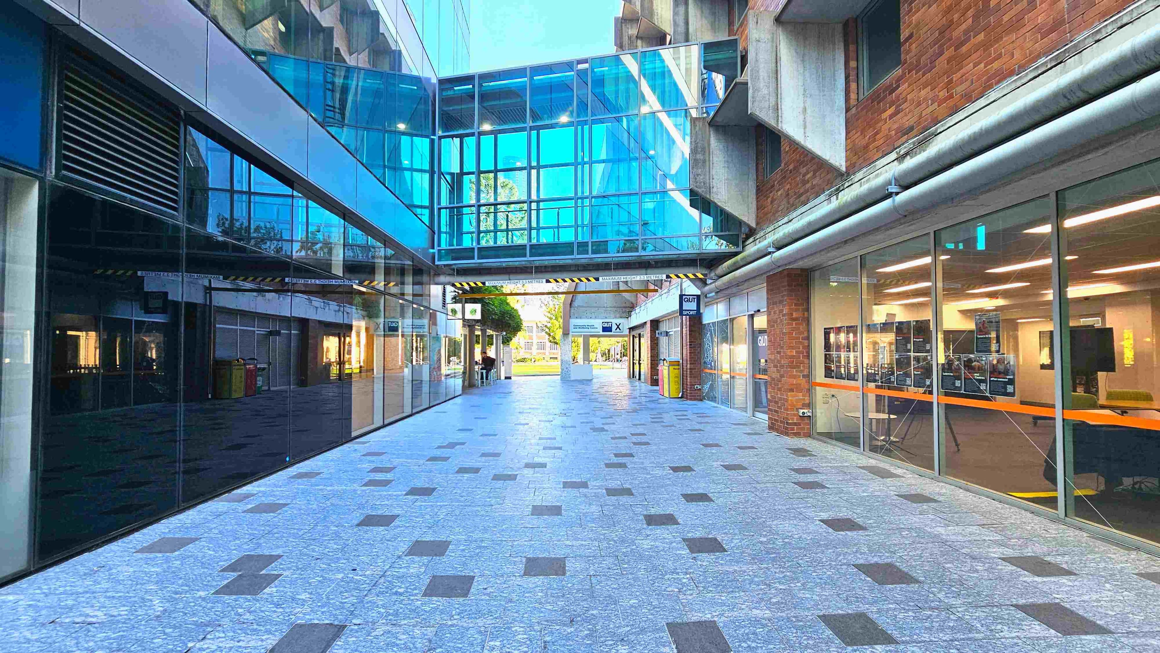 A modern walkway with a polished stone floor featuring square patterns, flanked by glass-fronted buildings. On the left, reflective glass panels mirror the scene, while the right side has brick walls and large windows displaying interior spaces. A blue-tinted glass skybridge connects the buildings overhead. Signs and bins are visible along the path, which leads to an open area with greenery in the distance.