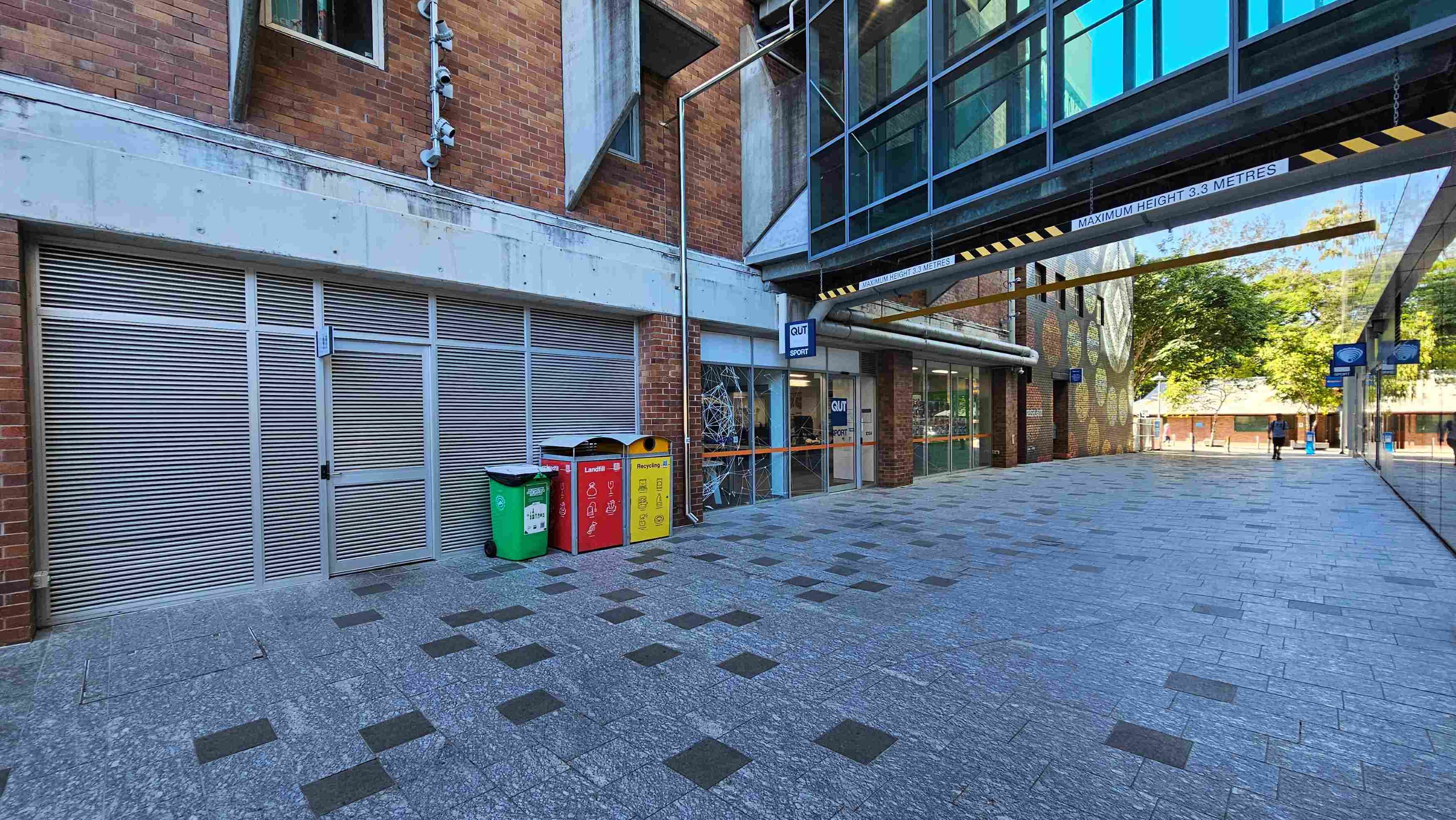 A modern urban walkway with a brick building on the left featuring metal louvred doors and a glass entrance labelled 'QUT Sport'. Three colourful bins for waste segregation are positioned nearby. Overhead, a glass bridge connects buildings, marked with a 'Maximum Height 3.3 Metres' sign. The ground is paved with grey tiles interspersed with darker squares. The area feels open, with trees visible in the background.