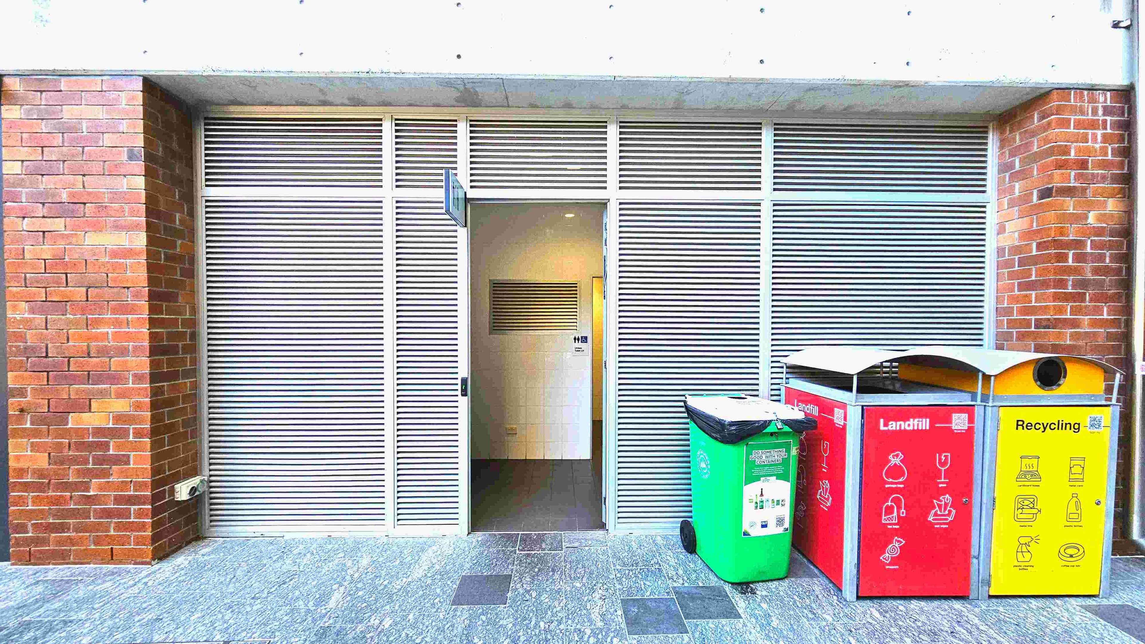 The image shows a public restroom entrance with a partially open door, set within a wall of horizontal metal slats. The entrance is flanked by red brick columns. To the right, there are three waste bins: a green one for general waste, a red one for landfill, and a yellow one for recycling. The floor is covered with grey stone tiles, some of which are darker, creating a subtle pattern.