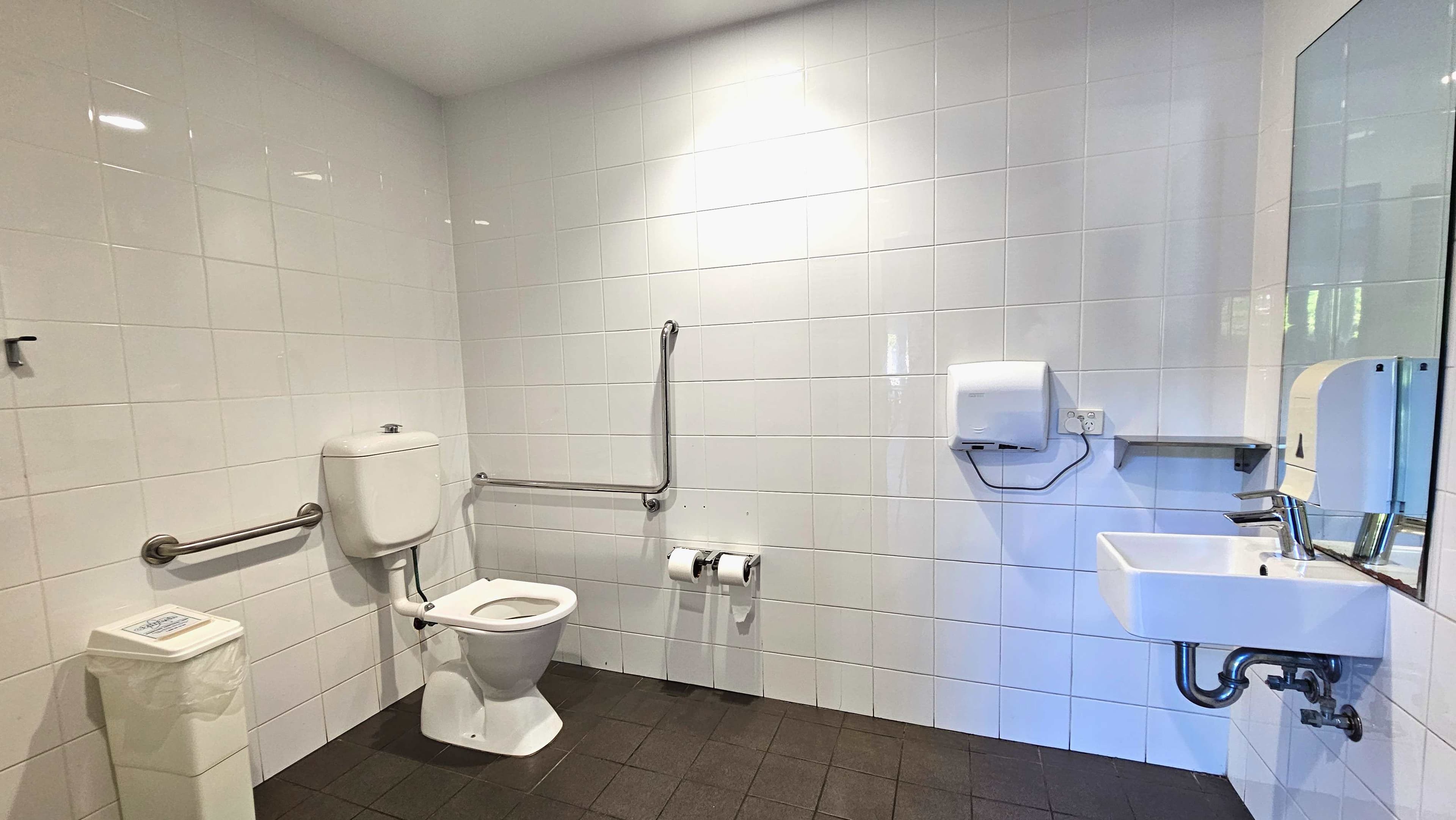 A clean, accessible bathroom with white tiled walls and a dark tiled floor. A toilet with a white cistern is positioned on the left, equipped with horizontal and vertical metal grab bars. A white bin is nearby. To the right, a wall-mounted sink with a mirror above it is visible, alongside a paper towel dispenser and a small shelf. Two toilet paper rolls are mounted on the wall between the toilet and sink.
