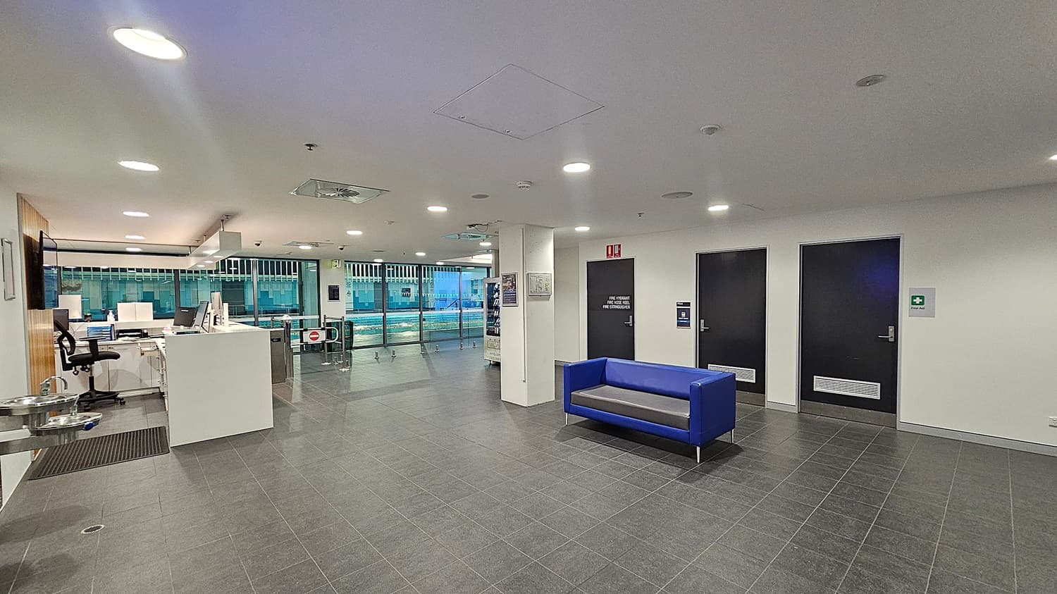 A modern indoor space with a grey tiled floor and white ceiling featuring recessed lighting. On the right, there are two black doors with signs, and a blue sofa against the wall. On the left, a reception area with a white desk, office chair, and computer equipment. A water fountain is mounted on the wall. In the background, large windows reveal a view of a swimming pool area with lanes and water.