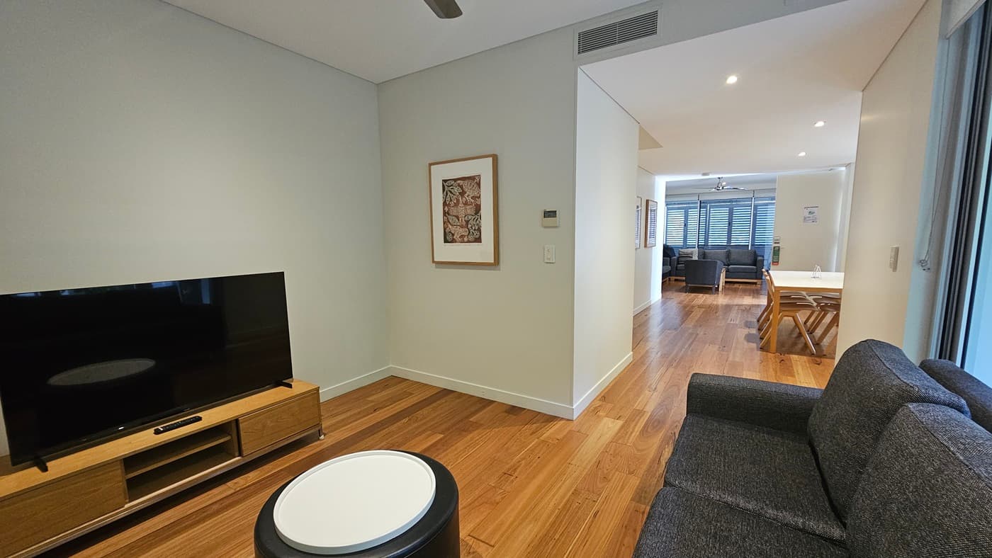 On the left, a flatscreen TV sits atop a TV stand. To the right, grey cushioned chairs are arranged against a glass wall. Further to the right, a dining table with wooden chairs and another lounge with grey cushioned chairs. The ceiling is all white with LED lights and a ceiling fan. The white wall features a framed decoration. The floor has timber designs.