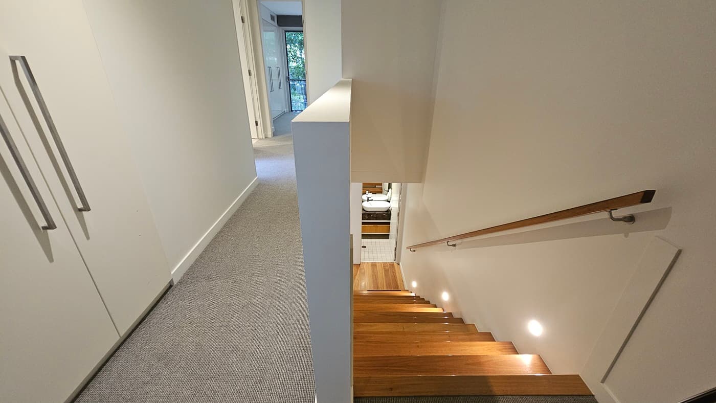 To the right is a wooden staircase with a wooden handrail leading to the upper level. The upper level features a carpeted floor. A white door with large handles is on the left. A wall partition secures the stairs. Further in, glass doors lead to the rooms.