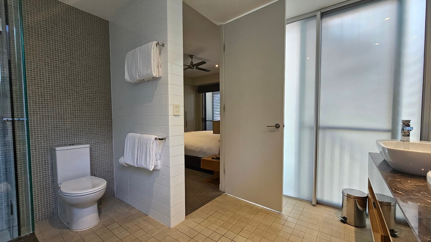 To the left, a toilet bowl is positioned against the grey wall. Towels hang on a handrail to the right. A glass wall is to its left, while a hand basin stands to the right, in front of a mirror. A rubbish bin is nearby. The wall on the right is made of glass and white and grey tiles in the middle. The floor has cream tiles. A section of the bedroom is visible.