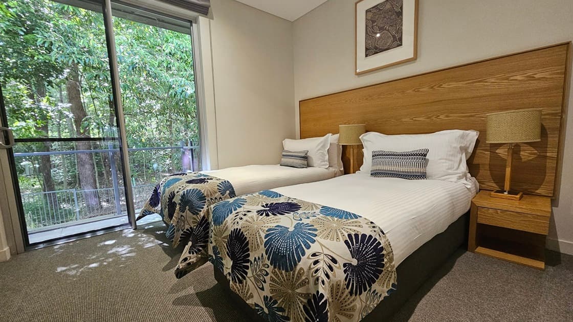 To the right, two single beds with a wooden headboard and matching bedside tables. Lampshades top the tables. To the left, retractable curtains on the left glass wall offer a scenic view of the greenery outside. A framed decoration hangs on top of the bed. The floor has grey carpet and the ceiling and walls are white.