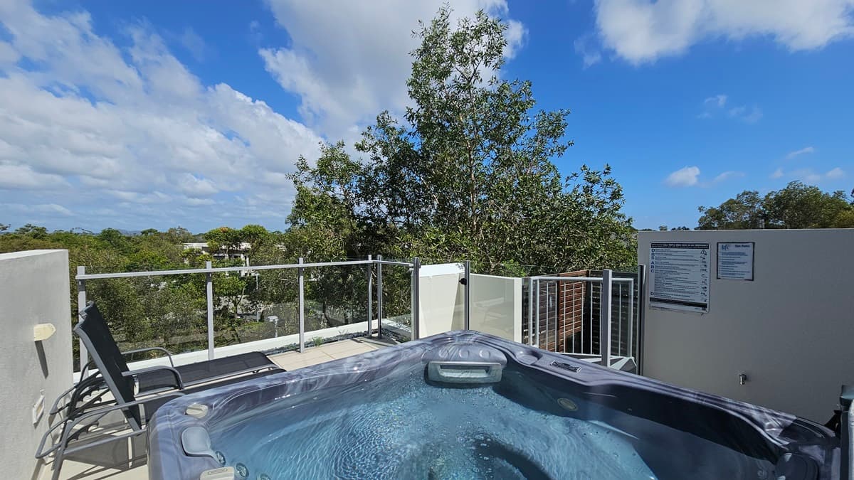 The rooftop spa features a grey hot tub in the middle, flanked by two black sun lounge chairs. Glass railings secure the area. Safety reminder posters are on the right wall. Trees surround the area.