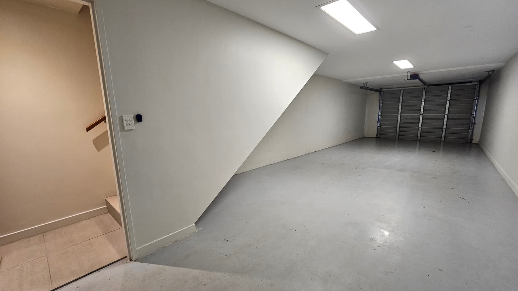 A space with a metal door and hard concrete floor. The white walls and ceiling are illuminated by fluorescent lights. A staircase with handrails on the left grants access to the upper level.