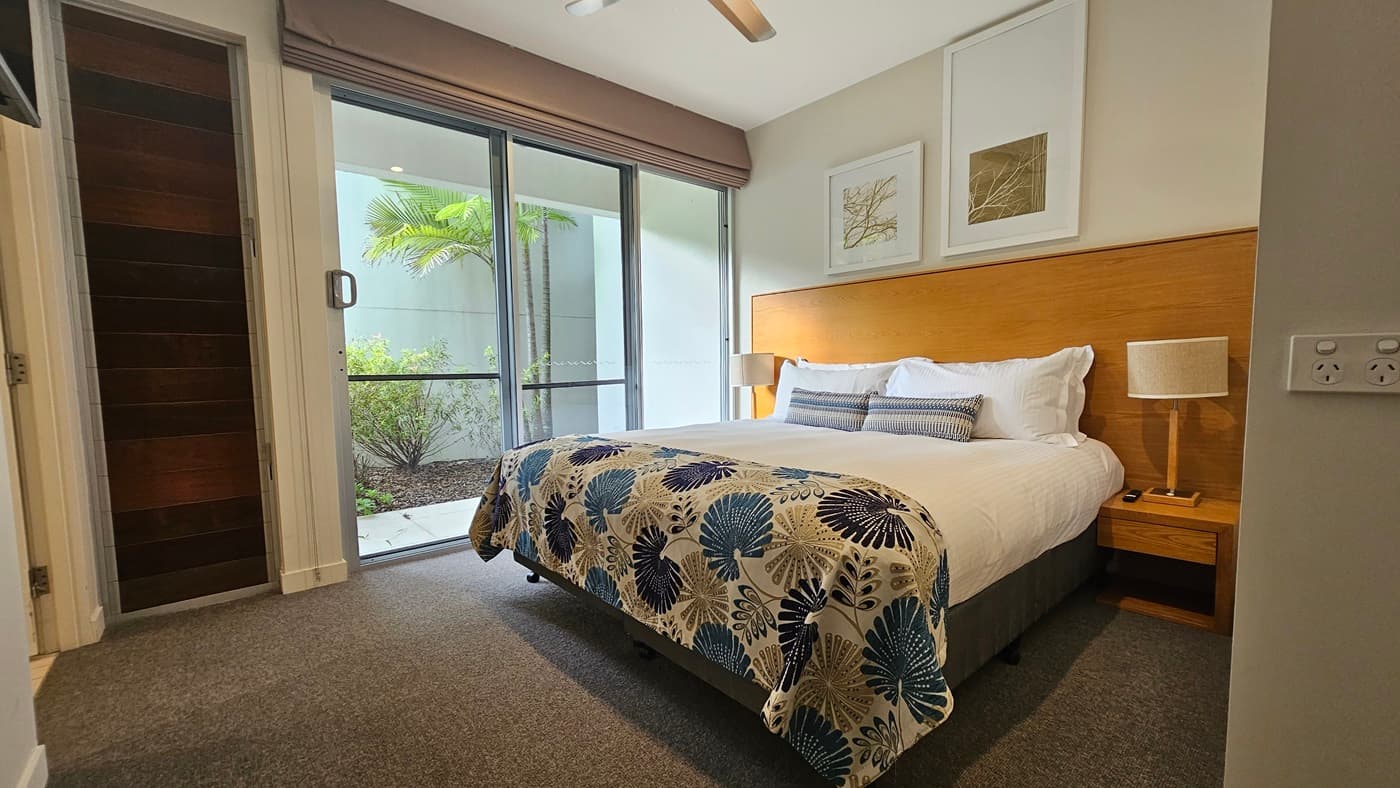 Inside the bedroom, a king bed with printed linens and a wooden headboard is flanked by matching bedside tables and lampshades. Large photo frames adorn the right wall. A glass door on the left leads outdoors, offering a view of small trees. A retractable curtain is affixed to the wall near a wooden door. The floor is covered in grey carpet.