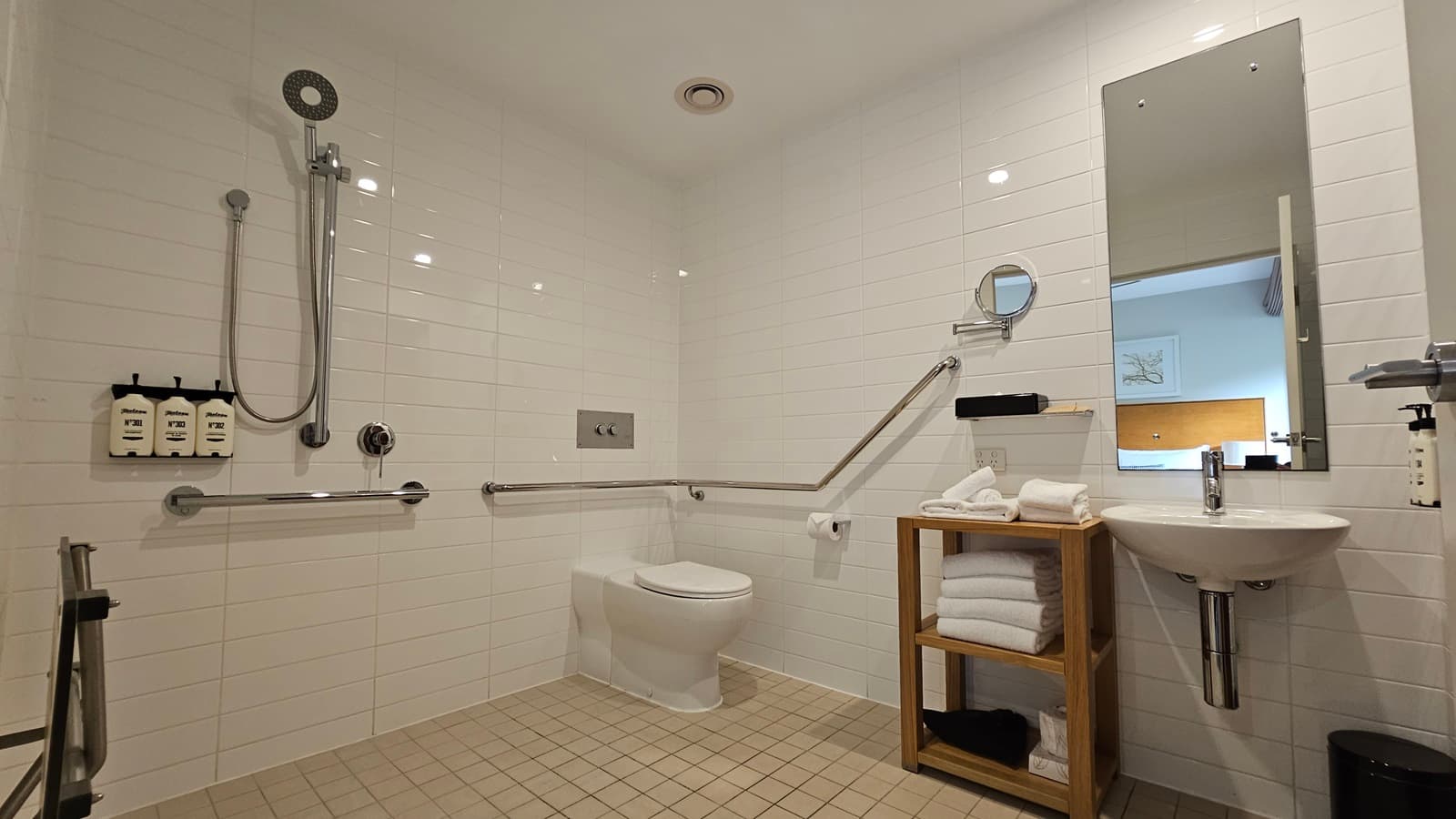 Inside the toilet, a toilet bowl with hand rails in the corner. Tissue dispensers are on the wall, with a shower and handrails to the left. Toiletries are affixed nearby. A wooden storage rack holds folded white towels on the right, beside a mirror, hand basin, and liquid soap dispenser. A rubbish bin is underneath. A foldable bench chair is near the shower. The floor has small cream tiles, while the walls and ceiling are white.