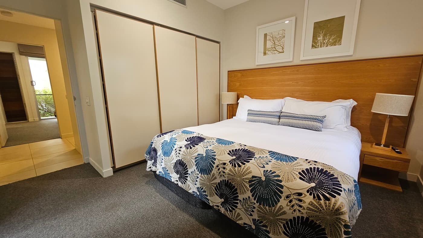 On the right is a king bed with printed linens and a wooden headboard, accompanied by matching bedside tables and lampshades. Large photo frames decorate the right wall. To the left is a wardrobe and a large mirror. The floor is carpeted in grey, while the ceiling and walls are white.