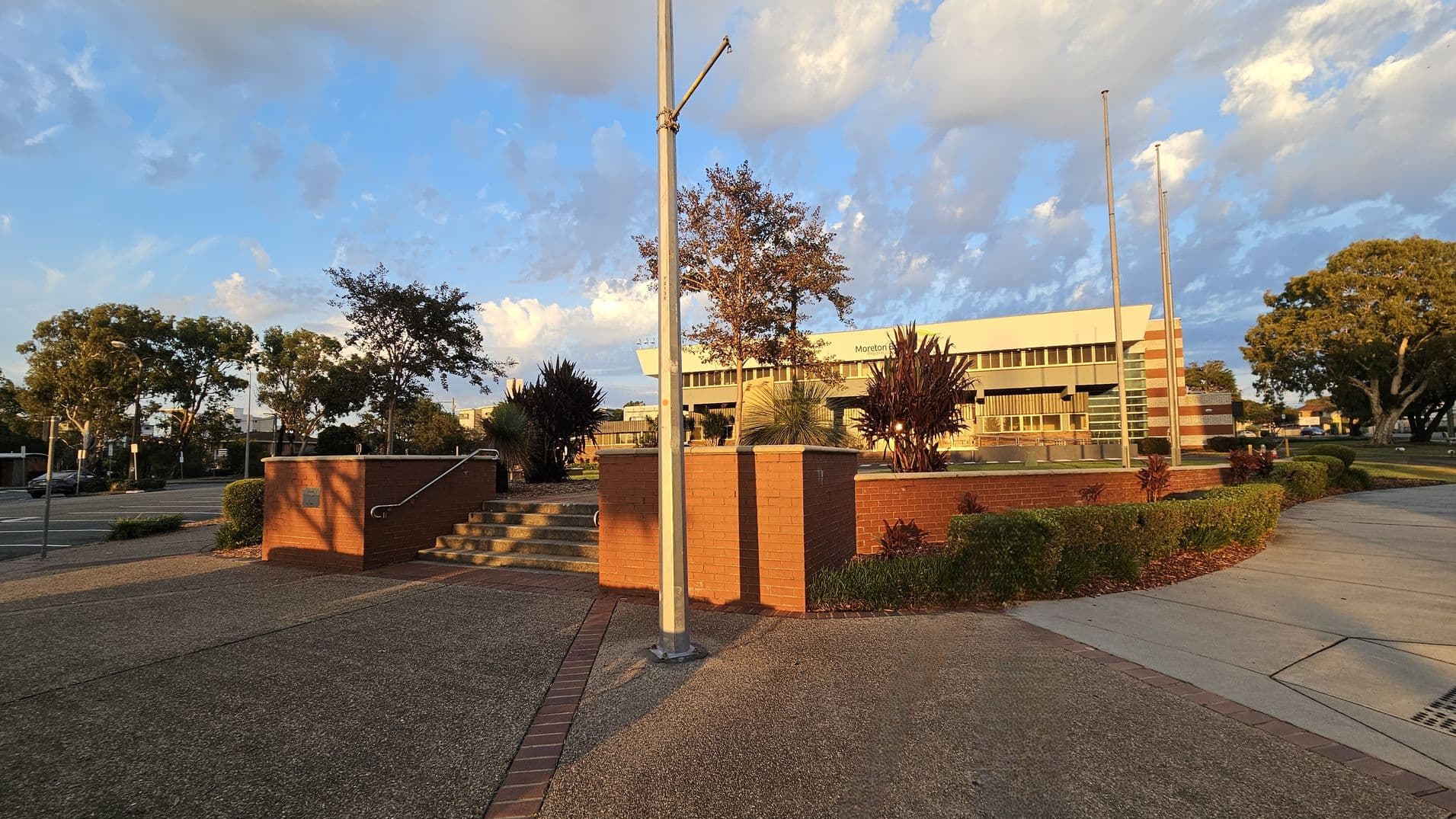 A paved area with a brick wall and steps in the foreground. A metal pole stands prominently in the centre. Behind, there are neatly trimmed bushes and small trees. In the background, a building with a sign that reads "Moreton" is visible, featuring large windows and a modern design. The area is surrounded by more trees and a road on the left. The sky is partly cloudy, casting shadows on the ground.