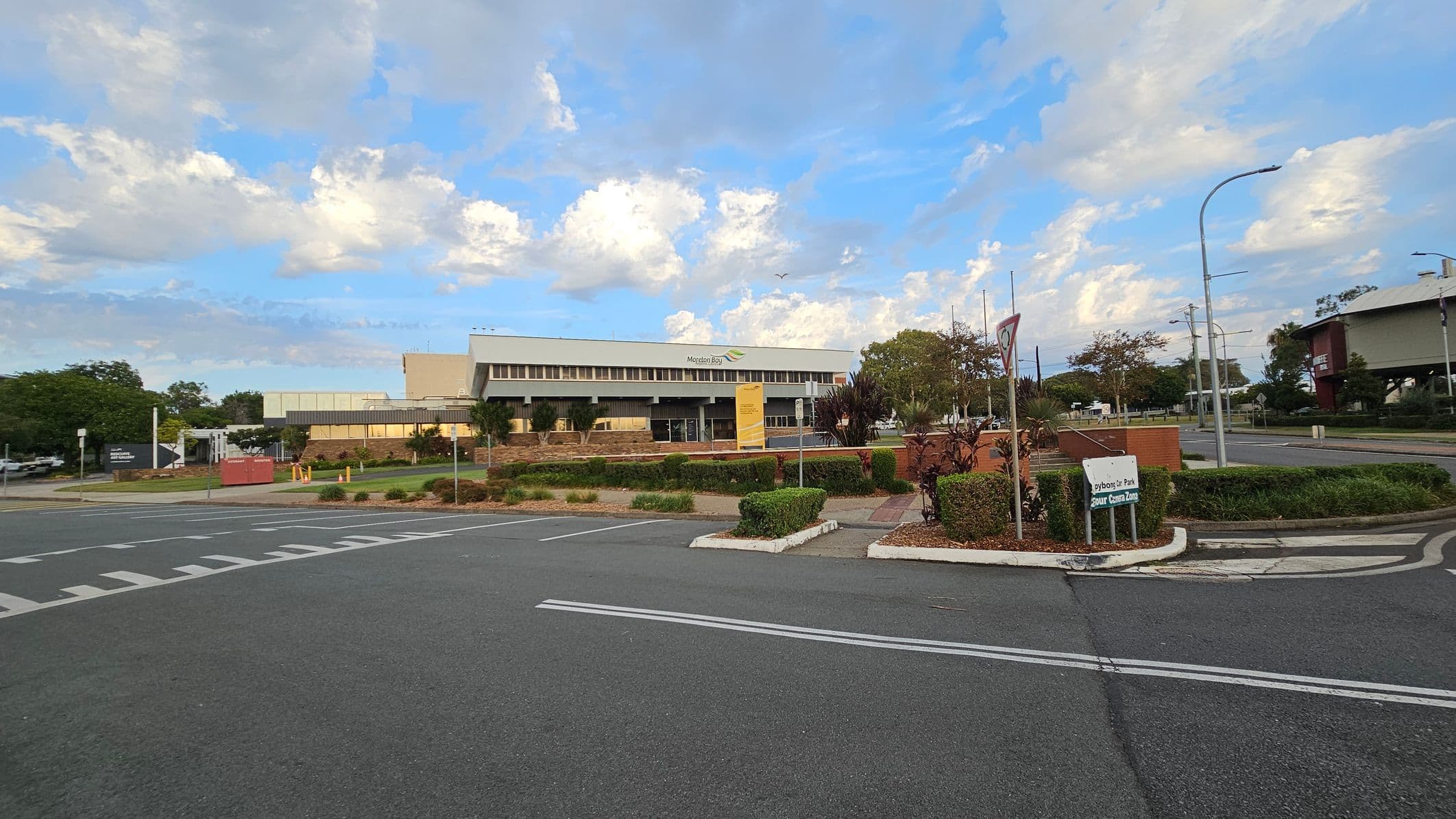 A large building with a modern design is in the centre, featuring a sign that reads "Moreton Bay." In the foreground, there is a paved road with white markings and a small roundabout. To the right, a sign indicates "Greyboys Park" and "Senior Citizens Zone." The area is landscaped with neatly trimmed bushes and small trees. On the left, there are more trees and a parking area. Streetlights and power lines are visible on the right side.