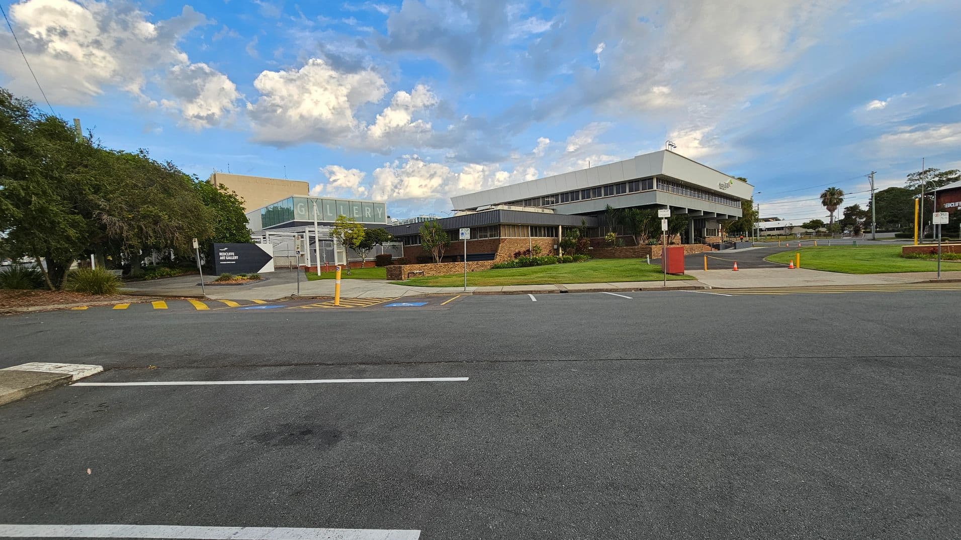 A wide street with parking spaces in the foreground. On the left, there are large trees and a sign pointing towards the "Art Gallery." The middle features a modern building with a glass facade displaying the word "GALLERY." On the right, a larger building with a flat roof and multiple windows. The area is surrounded by well-maintained grass and a few small trees. Orange traffic cones are placed near the entrance of the building.