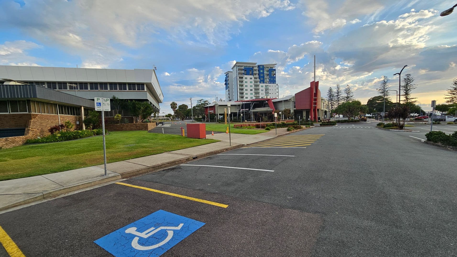 A parking area with a marked disabled parking space in the foreground. On the left, there's a brick building with a sloped roof and a grassy area with a signpost. In the centre, a tall building with blue and white sections is visible. On the right, a modern building with red accents and a row of trees along the street. The road has yellow lines and a few parked cars are visible in the distance.