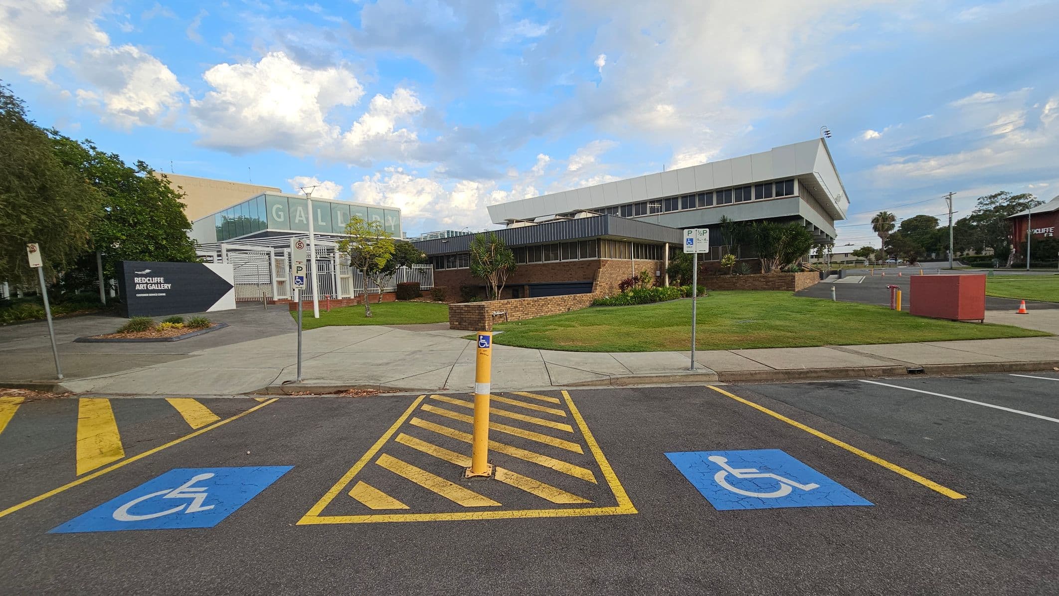 In the foreground, there are two parking spaces marked with blue wheelchair symbols and yellow lines. A yellow bollard is positioned between them. The background features a modern building with a sign for the Redcliffe Art Gallery on the left. The building has a mix of glass and brick elements, with a grassy area and small trees in front. A paved walkway leads to the entrance. The sky is partly cloudy.