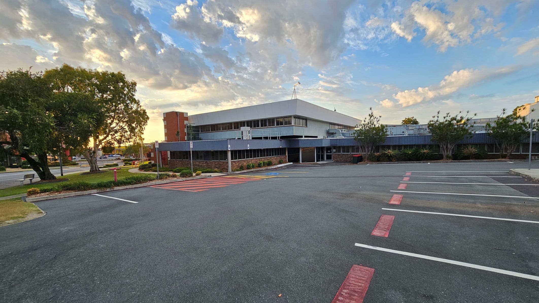 A large, modern building with a flat roof is in the centre, featuring a mix of glass and metal panelling. In the foreground, there is an empty car park with marked spaces and red pedestrian crossings. On the left, there are several large trees and a grassy area with a bench. To the right, smaller trees and shrubs line the building. The background shows a few more buildings and a road with light traffic.